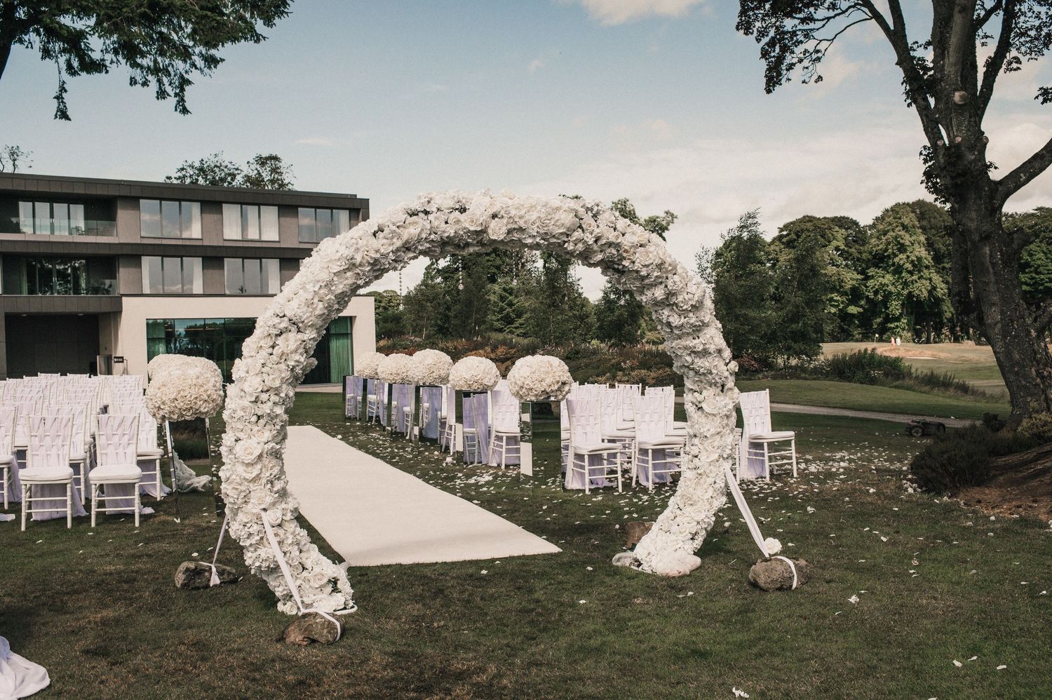 Elegant white floral wedding arch frames an outdoor ceremony space with white chairs.