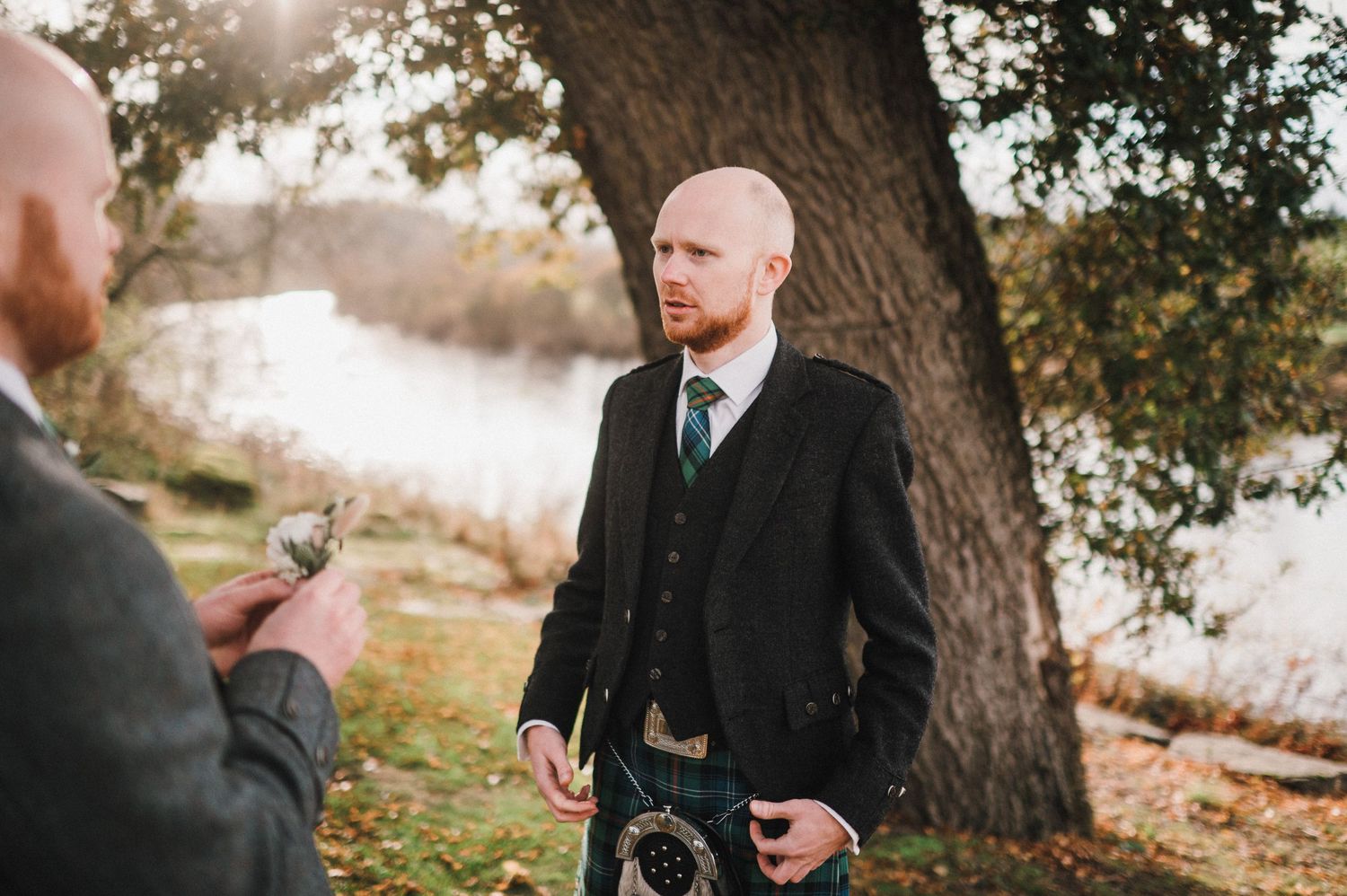 Person in formal wear and kilt standing by tree near water while being photographed.