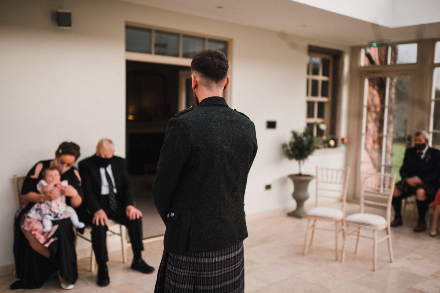 Person in traditional Scottish kilt photographing seated guests at an event venue.