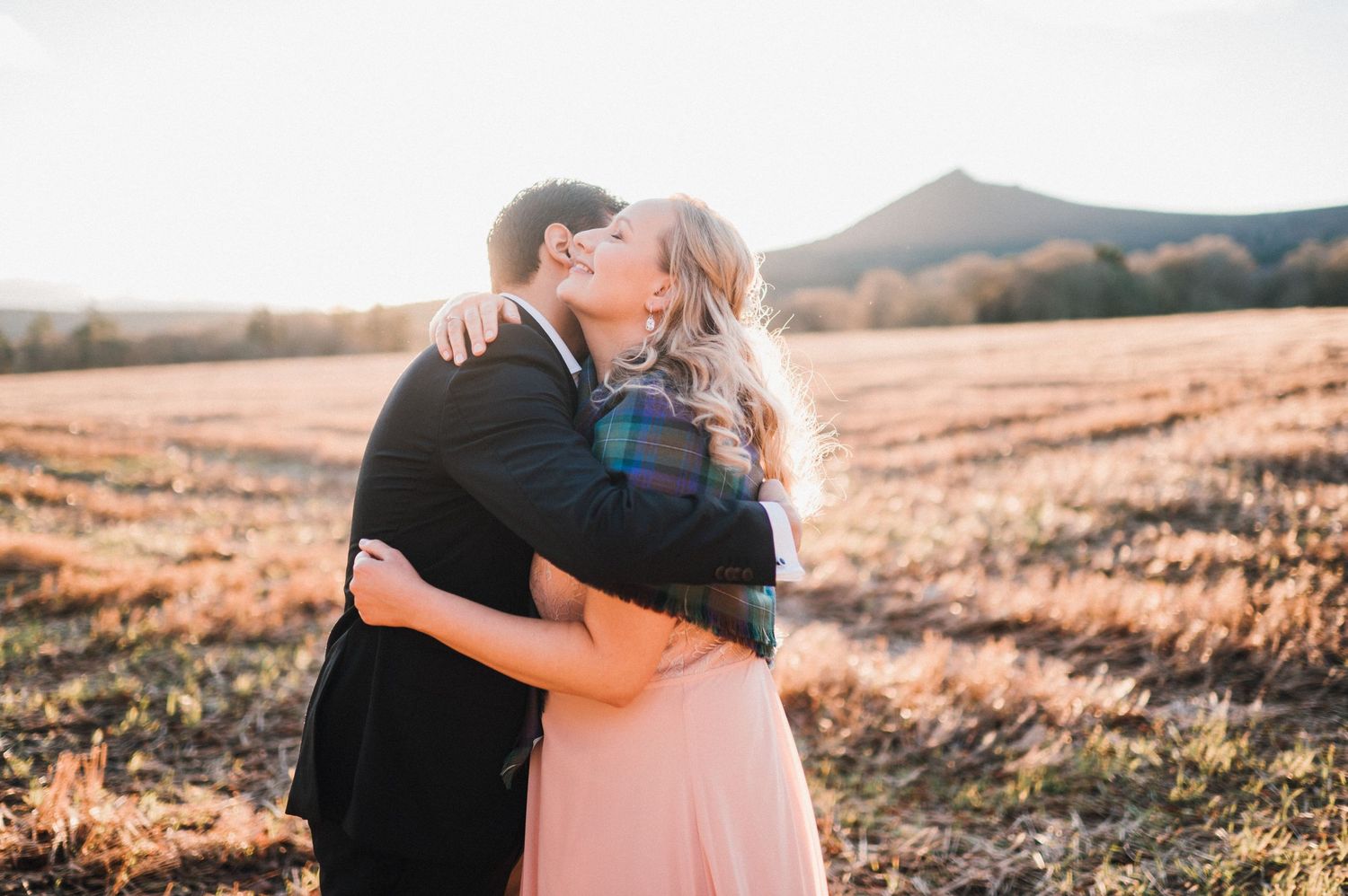 A couple embraces in golden sunlight with rolling hills behind them.