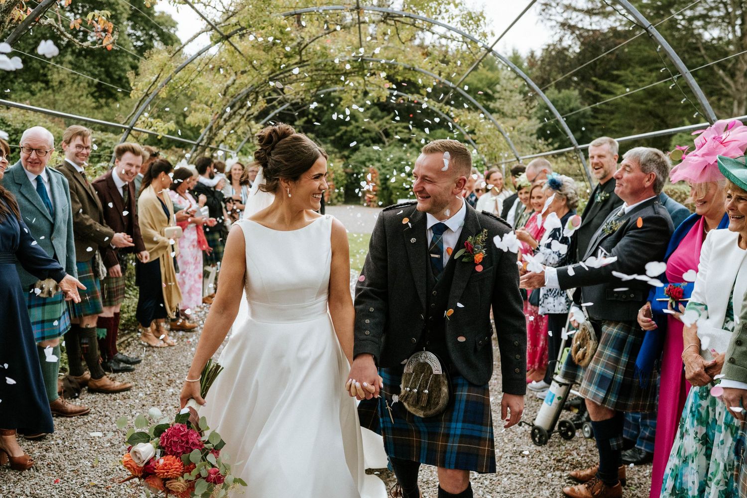 Wedding celebration with guests in colorful attire and kilts gathered on a garden path.
