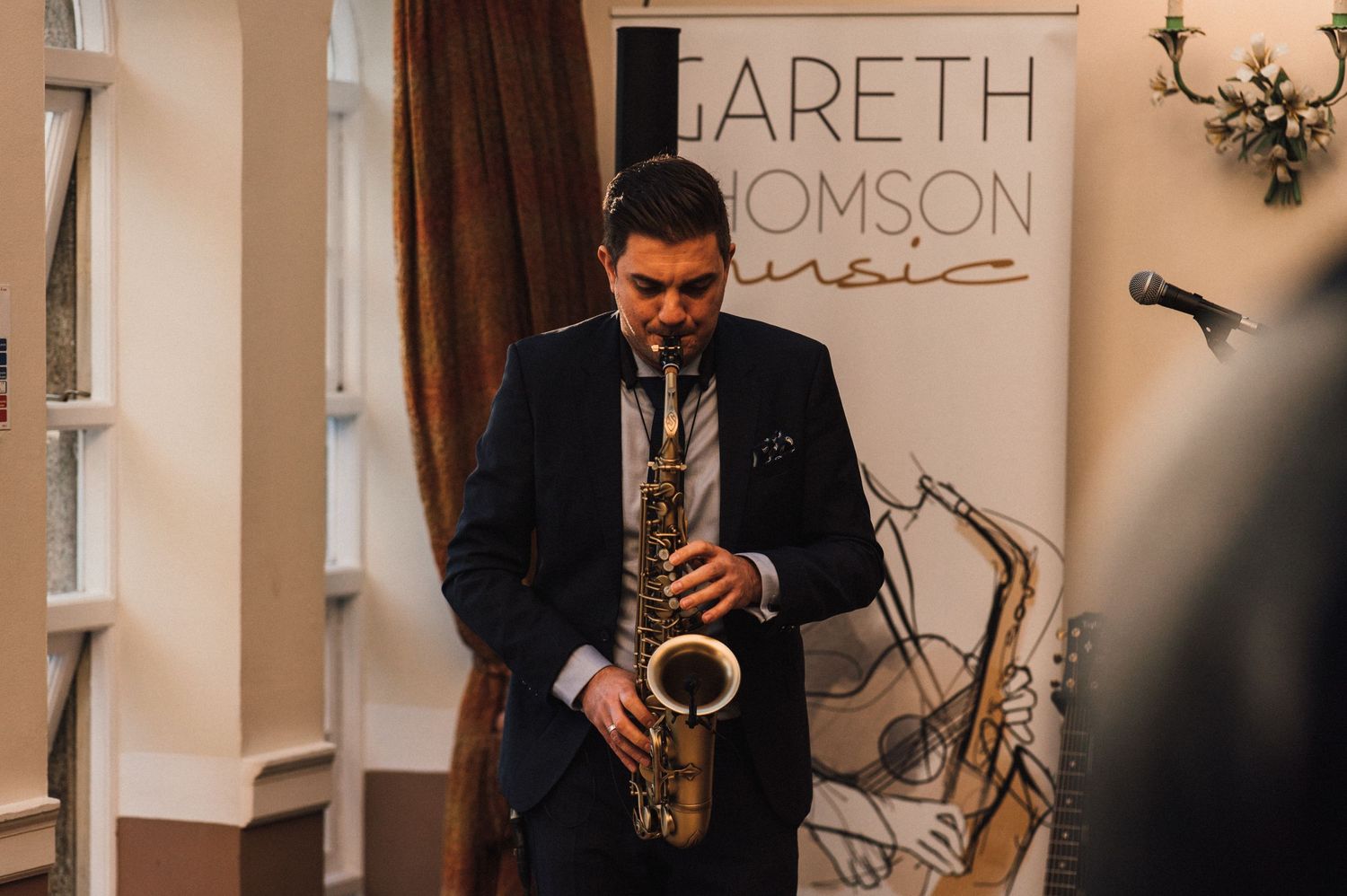 A musician plays saxophone in front of a branded backdrop at an event venue.