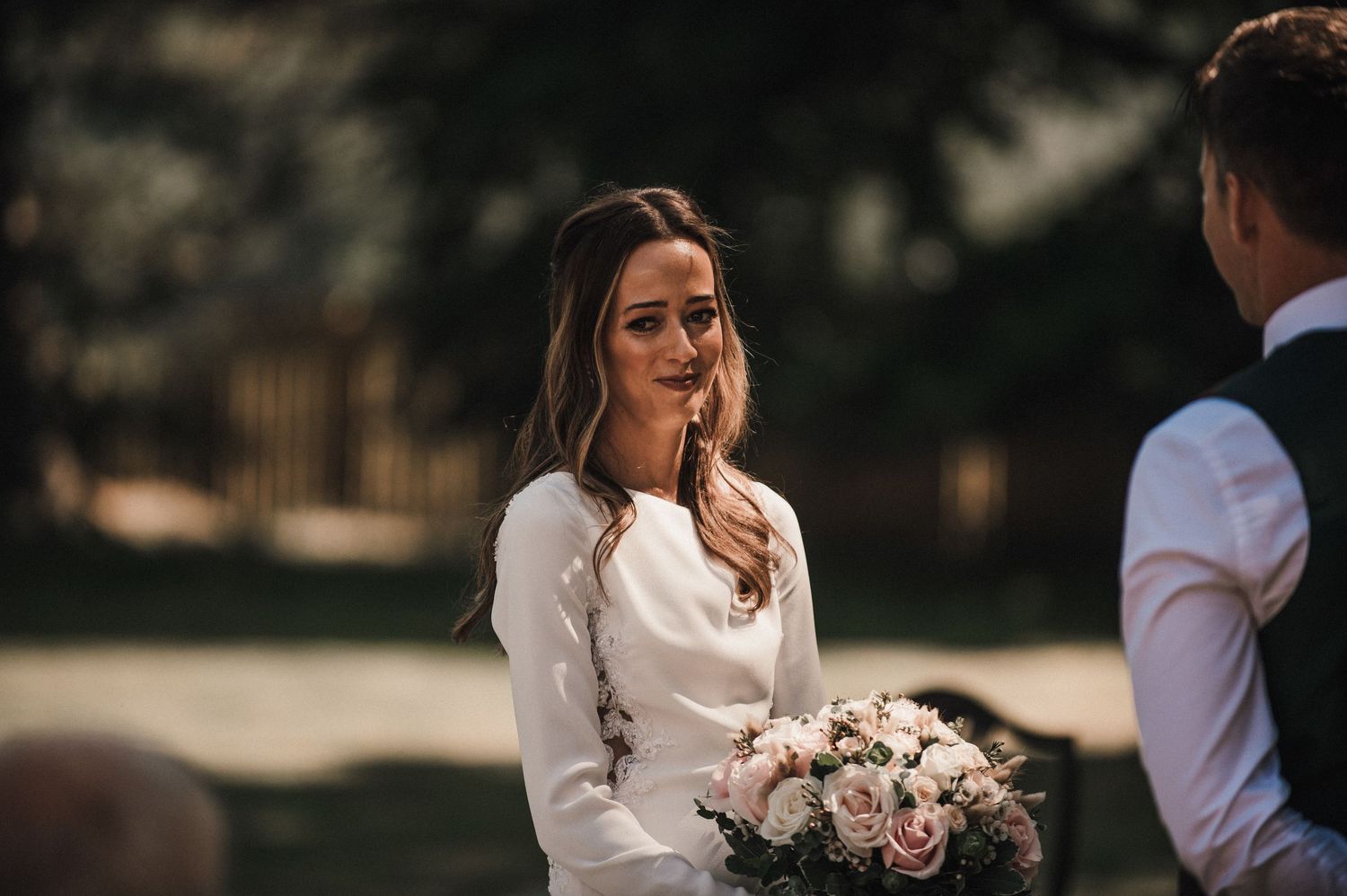 A bride in a sleek white dress holds a pink and white bouquet during an outdoor ceremony.
