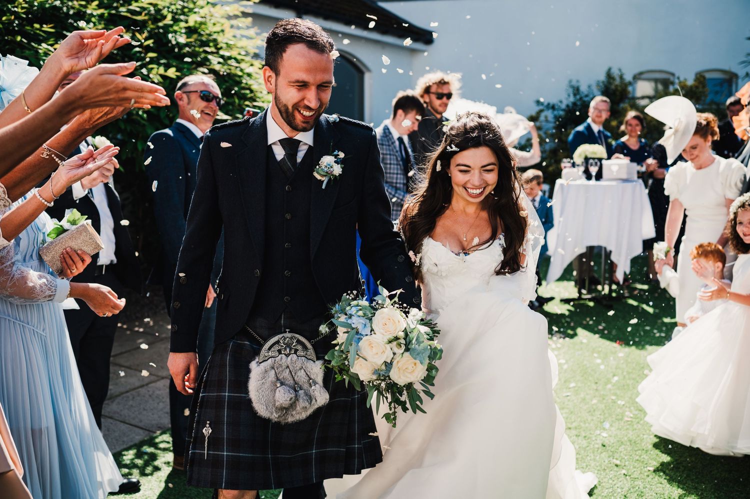 Wedding couple walks down a path surrounded by guests while confetti falls on a sunny outdoor ceremony.
