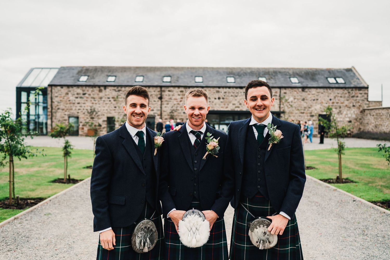 Three men in formal kilts and suits pose in front of a stone building.