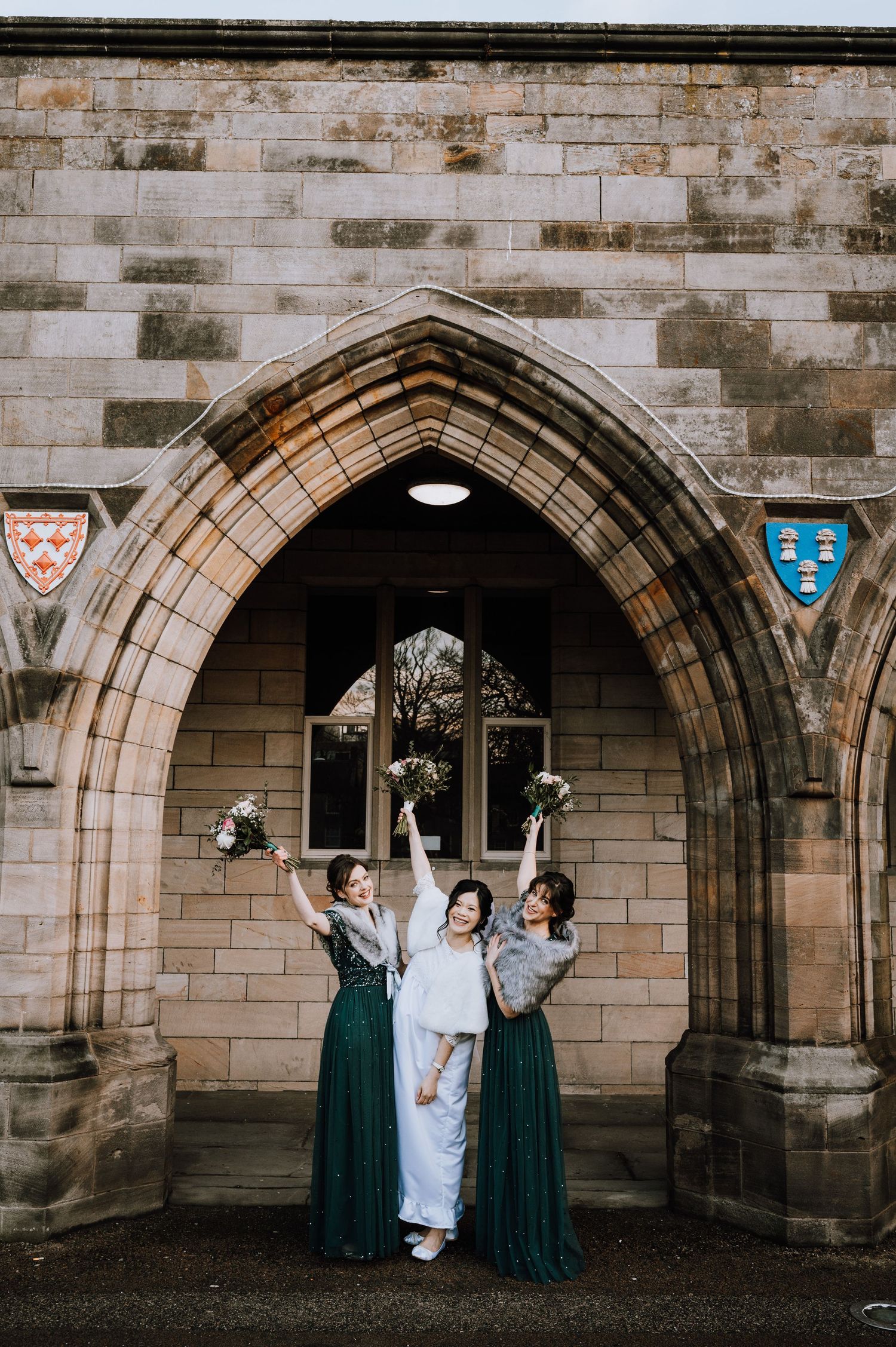 Bridesmaids in emerald green dresses celebrate with bride under Gothic stone archway at church wedding.
