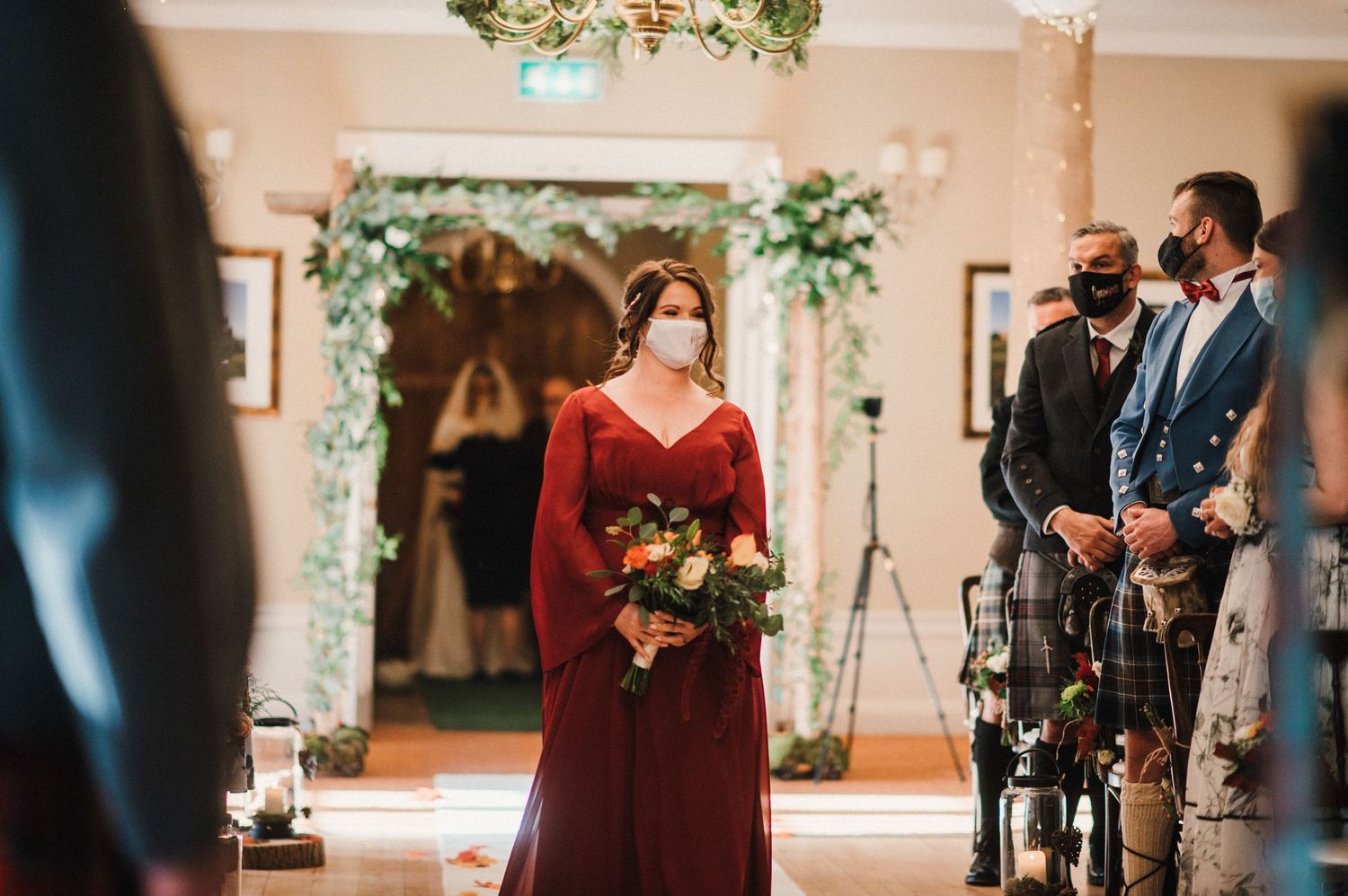 Bridesmaid processes down aisle in burgundy gown beneath floral arch decorations.