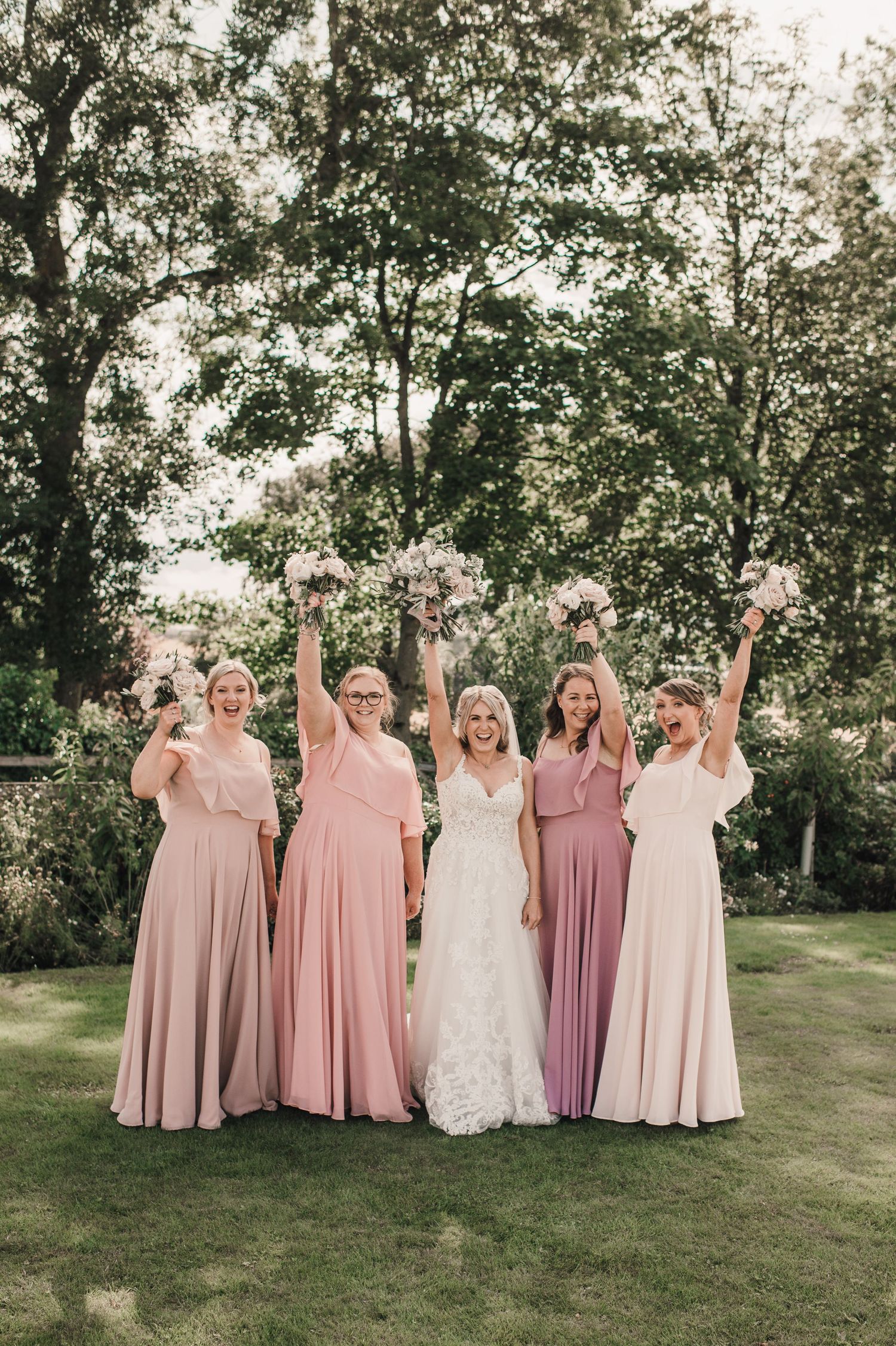 Wedding party in pink and blush dresses poses with raised bouquets in garden setting.