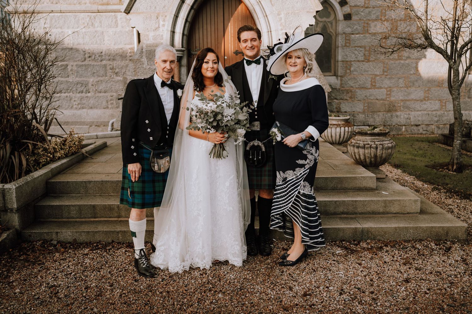Small wedding group in Scottish formal wear and kilts pose on stone church steps.