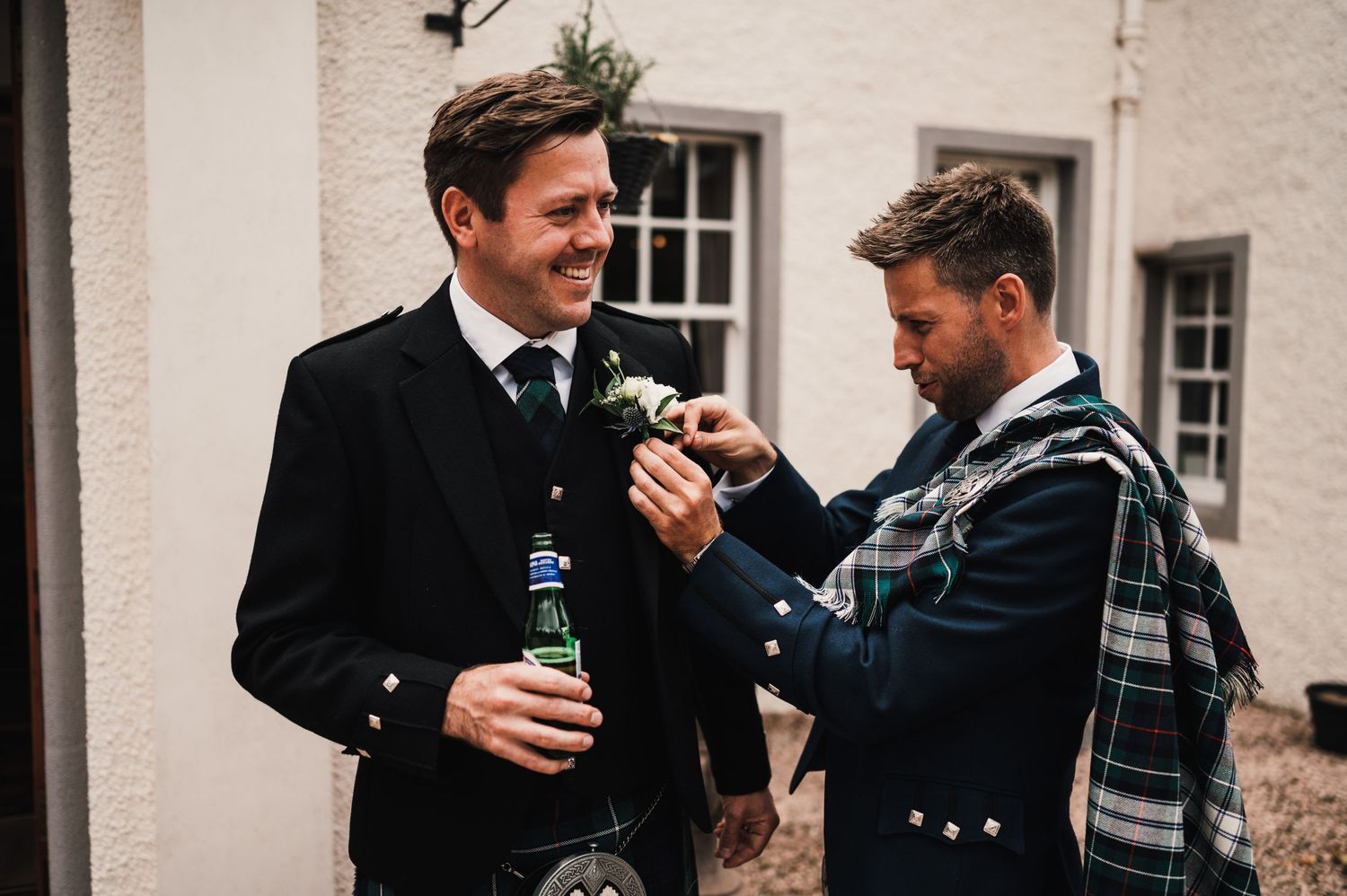 Two men in Scottish formal wear sharing drinks and conversation outside a white building.