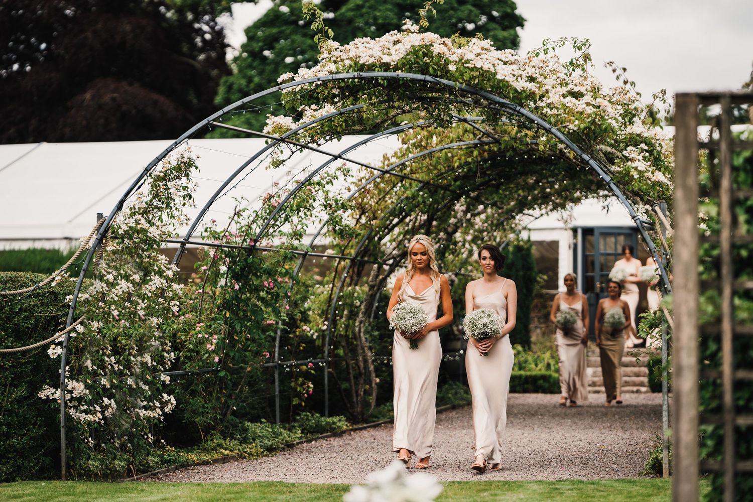 Two people in formal attire walk under floral archway on garden path.