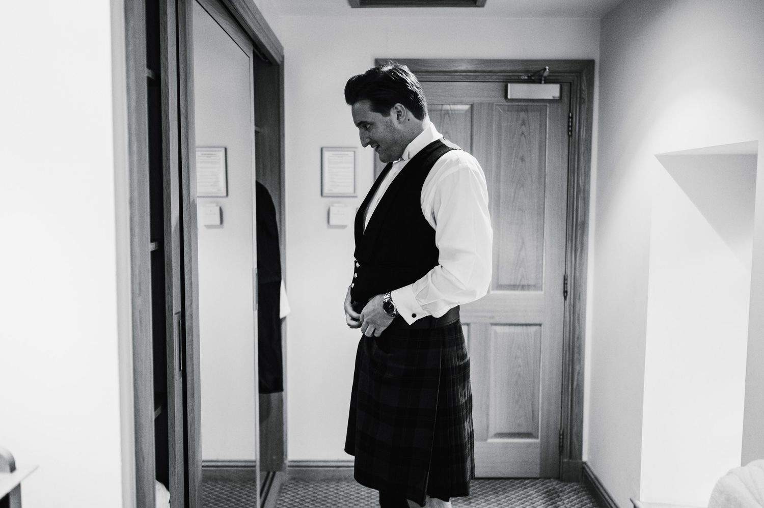 Black and white image of formal Scottish attire preparation in a hotel hallway.