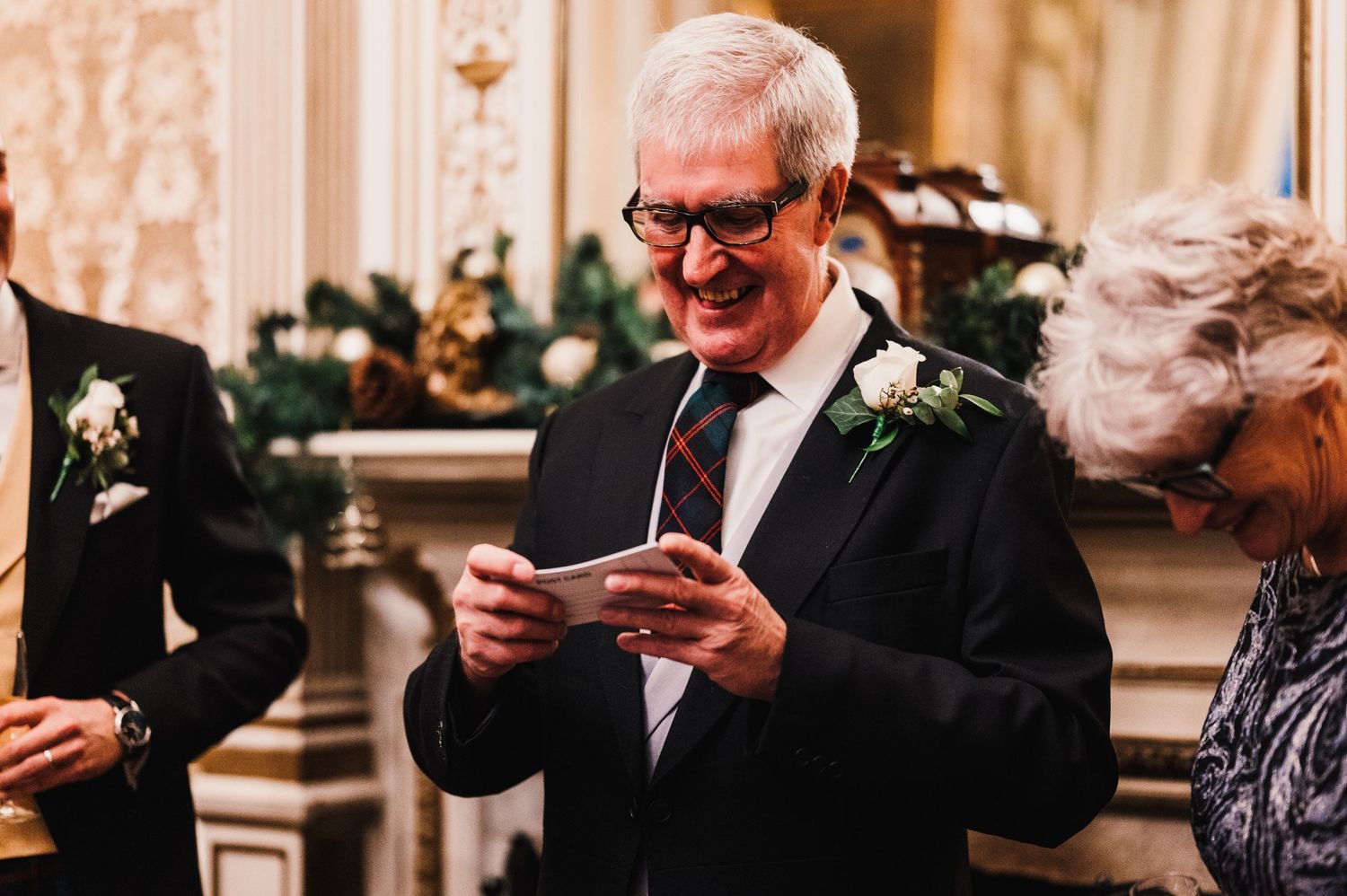 A gentleman in formal attire reads from a card during an elegant indoor wedding celebration.