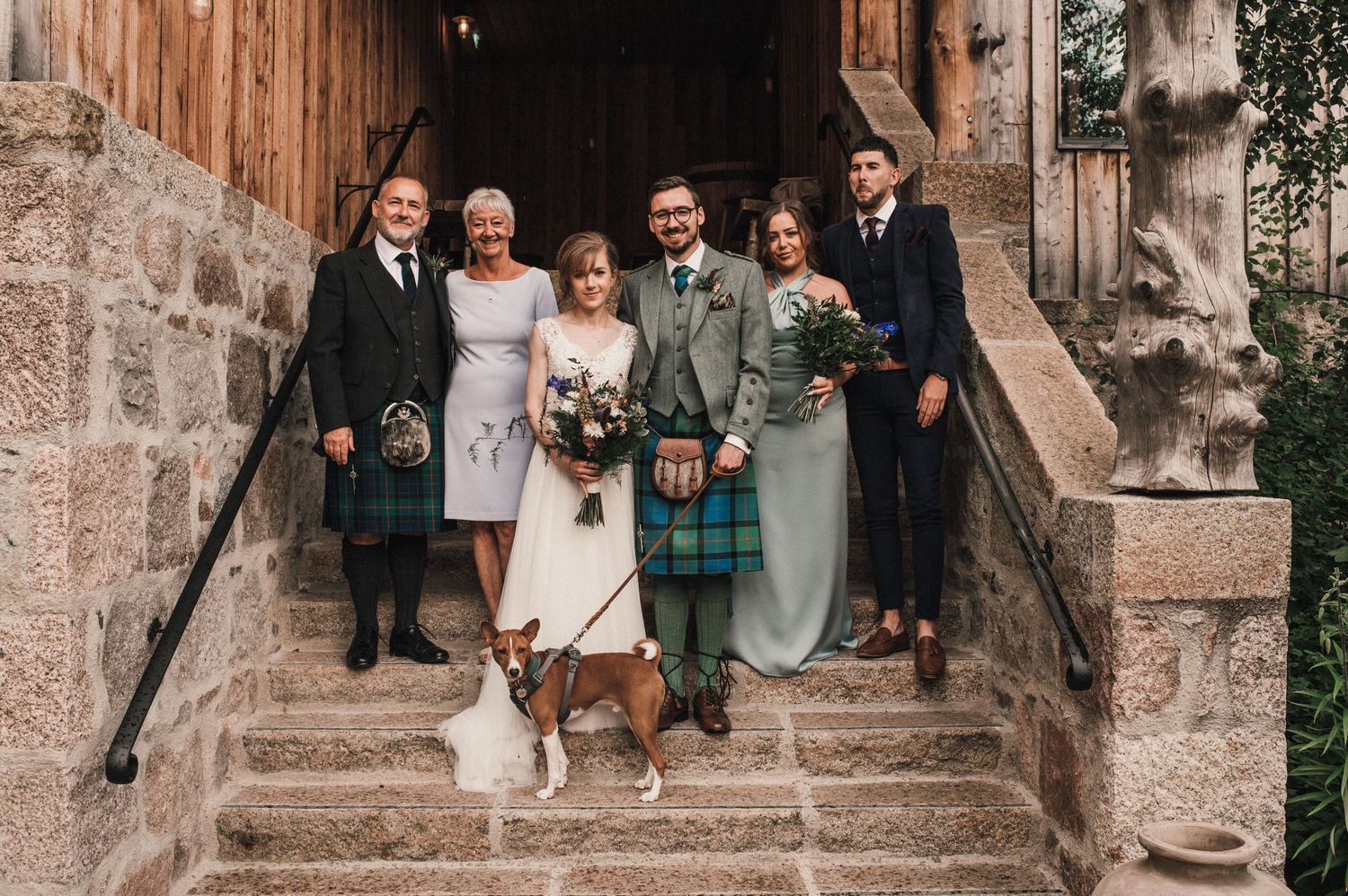 Small wedding group poses on stone steps with dogs in a rustic venue.