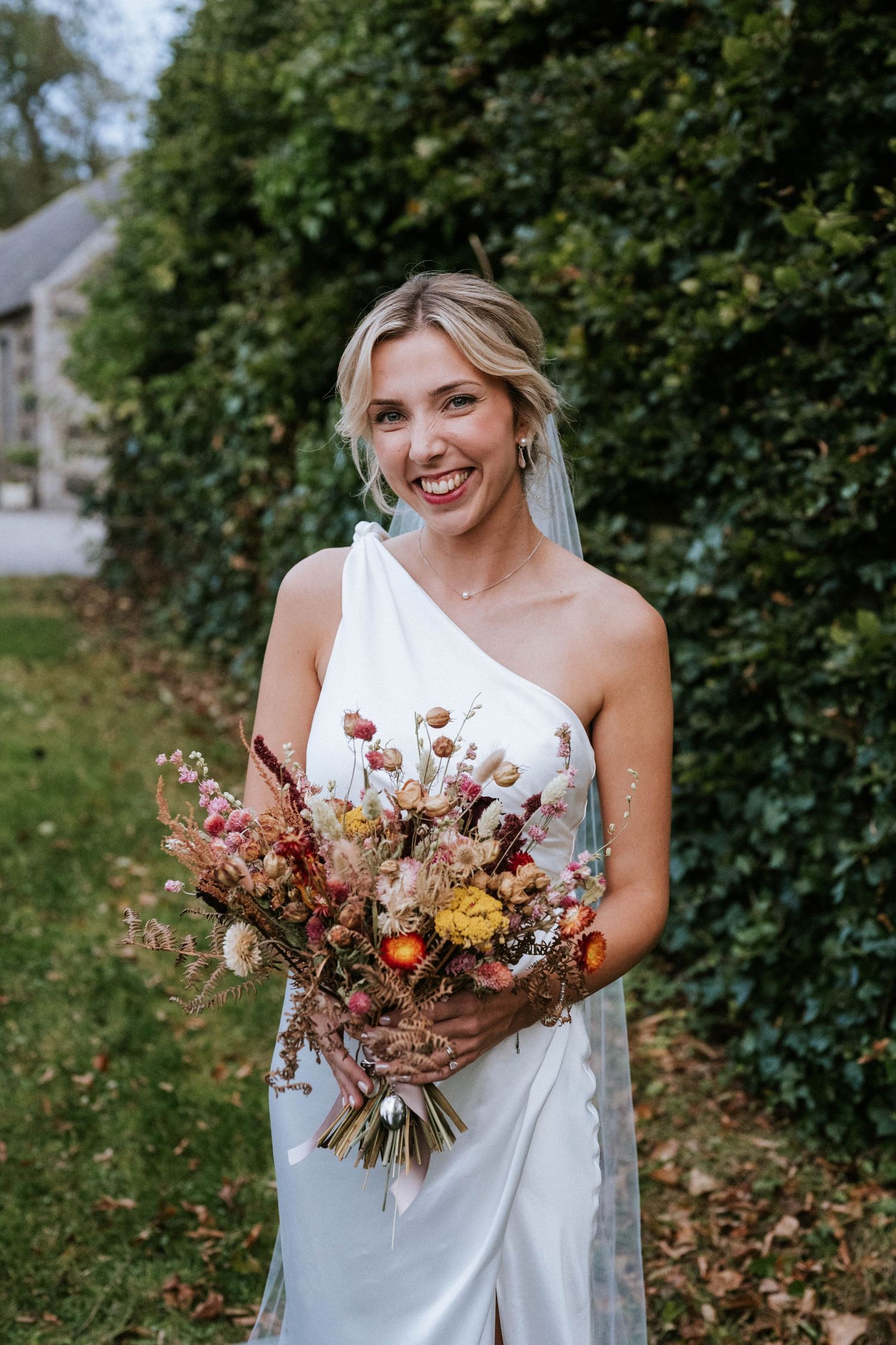 A one-shoulder white dress adorned with autumn-toned dried flower bouquet in garden setting.