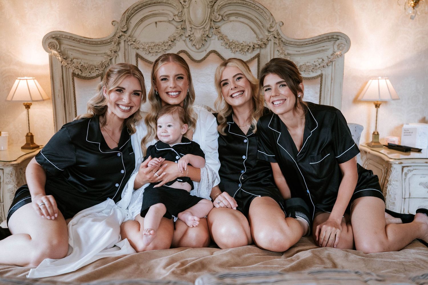 Group wearing matching black robes lounges together on white bedding for casual pre-wedding photos.