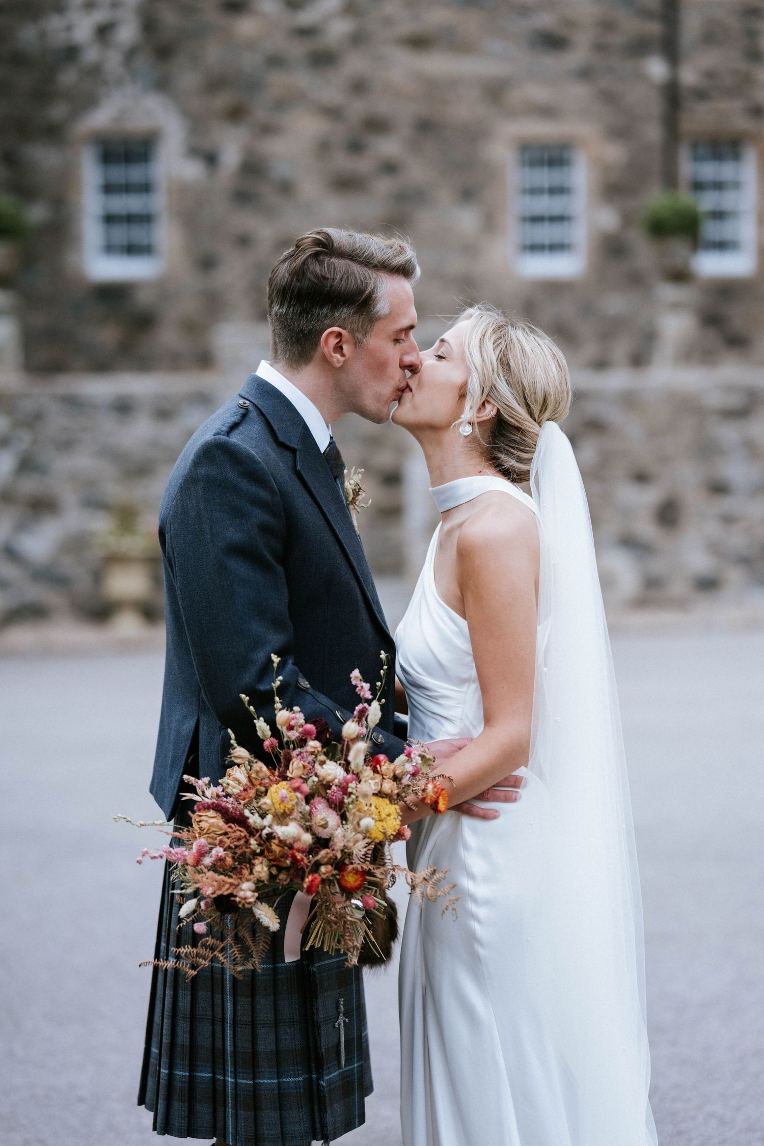 Romantic wedding portraits with the groom in traditional Scottish kilt outside a stone building.