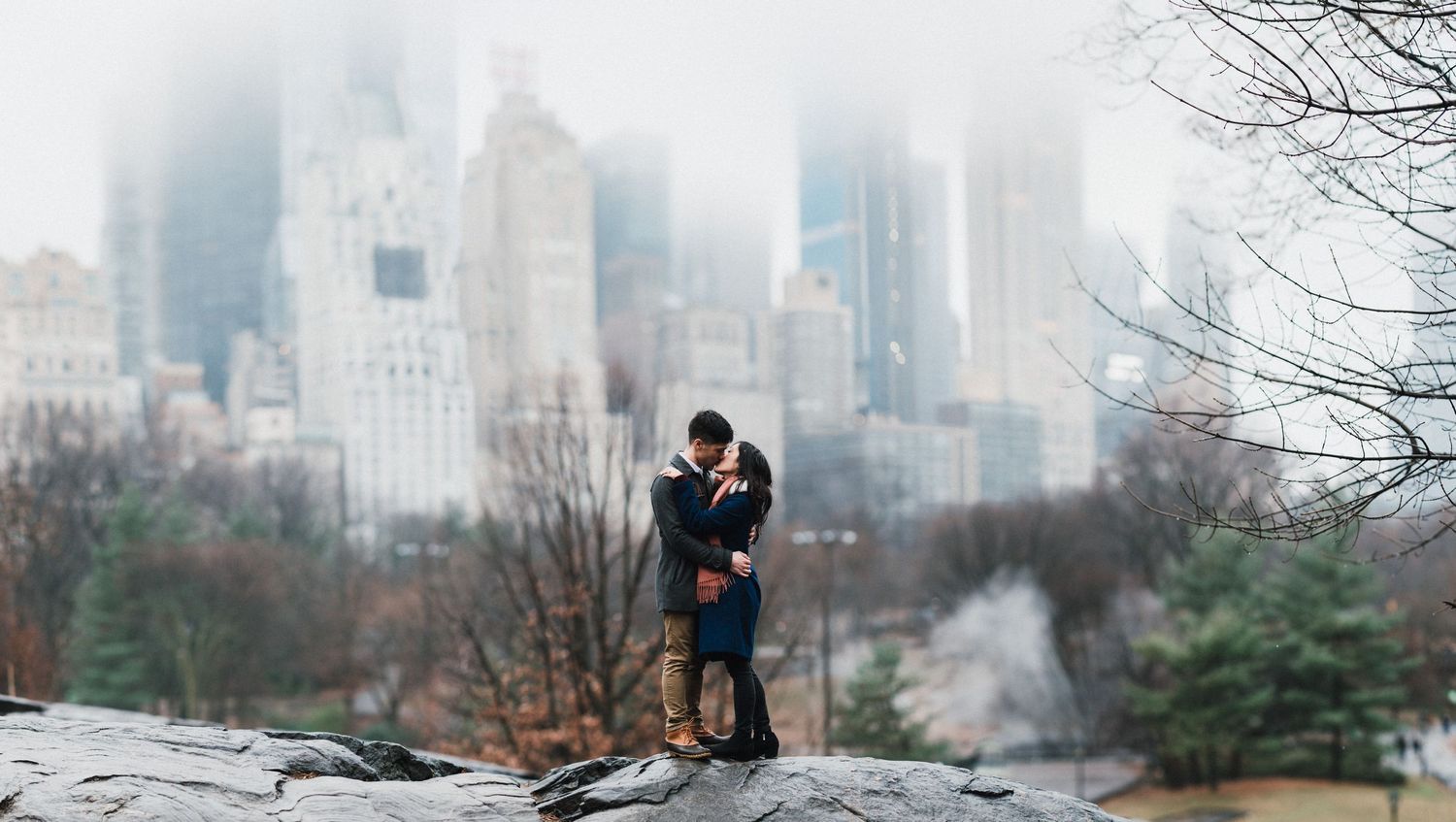 A couple shares an intimate moment on a rocky outcrop overlooking a misty Central Park with Manhattan skyline in the background.