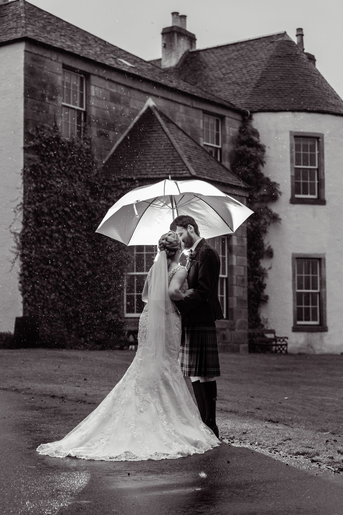 A romantic wedding couple shares an umbrella in the rain outside a Scottish stone manor house.