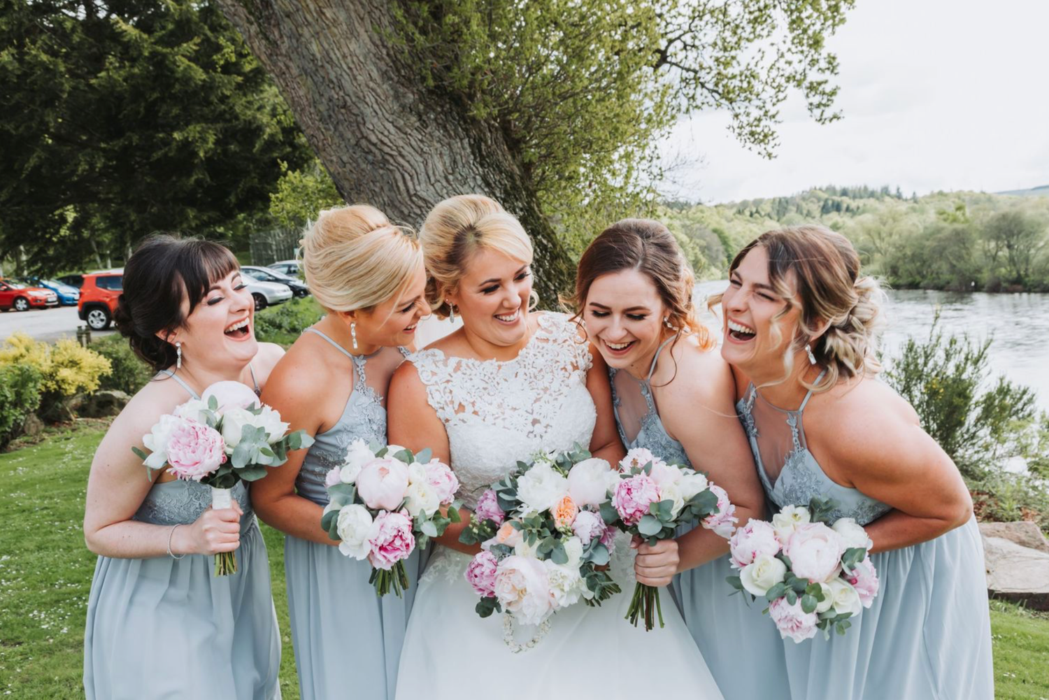 A group of bridesmaids in light blue dresses hold pink and white bouquets while laughing together outdoors.