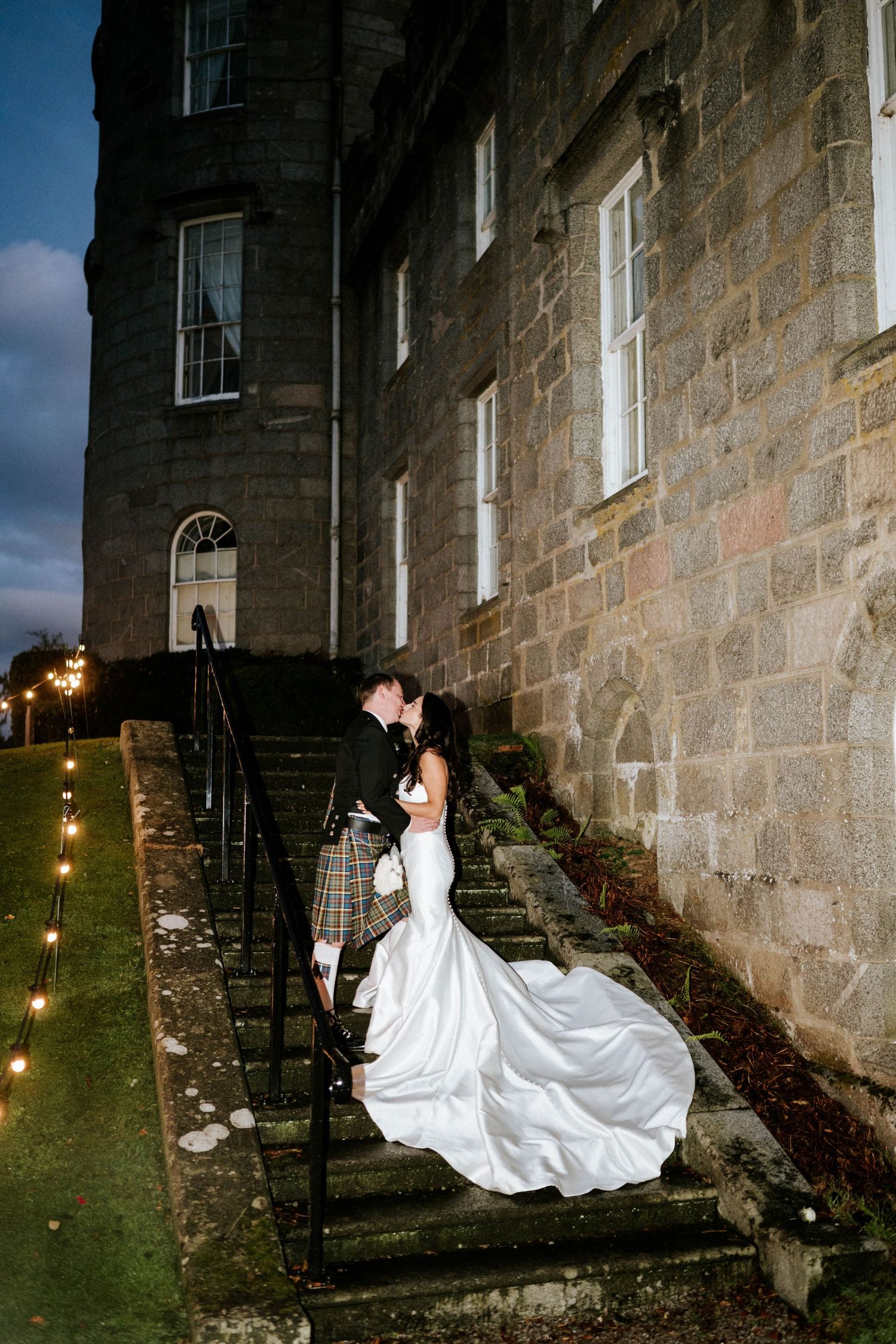 A wedding couple poses on stone steps of a historic building with string lights creating ambiance.