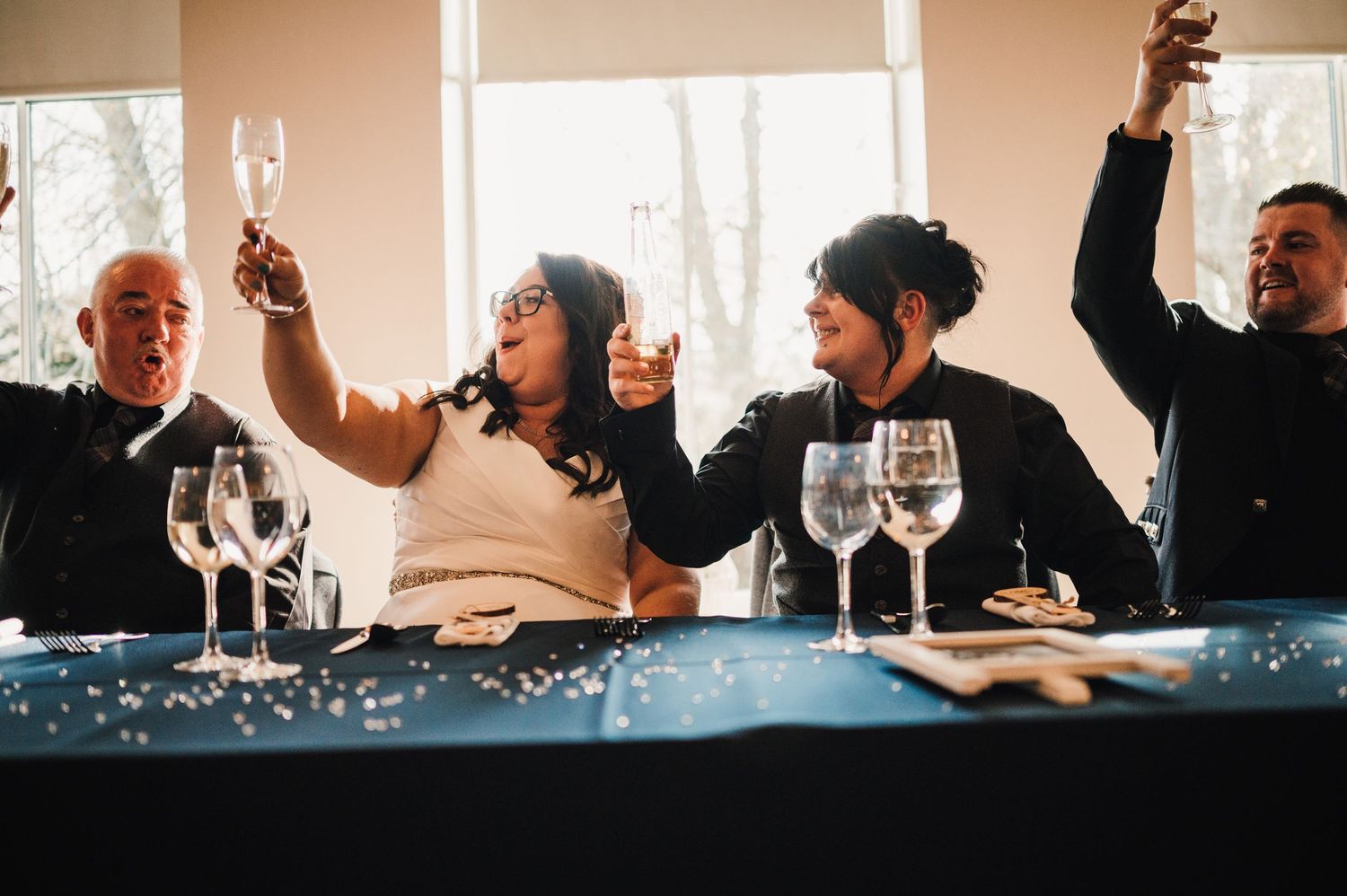 Logie House Wedding Photography. Joyful wedding guests celebrating with raised glasses at reception table.