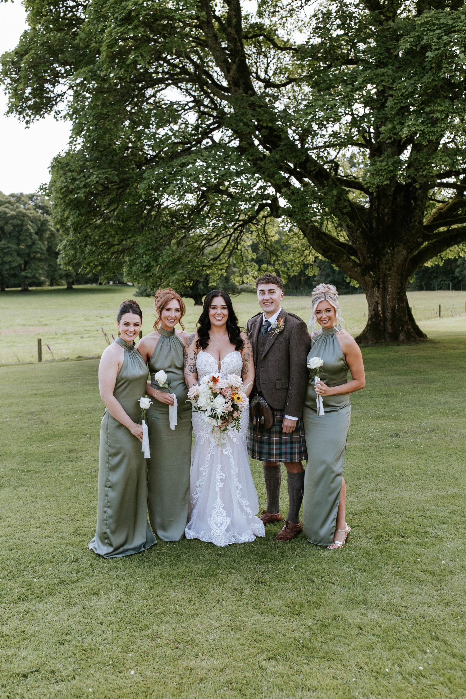 Wedding group photo featuring sage green bridesmaid dresses and traditional Scottish kilt under a majestic tree.