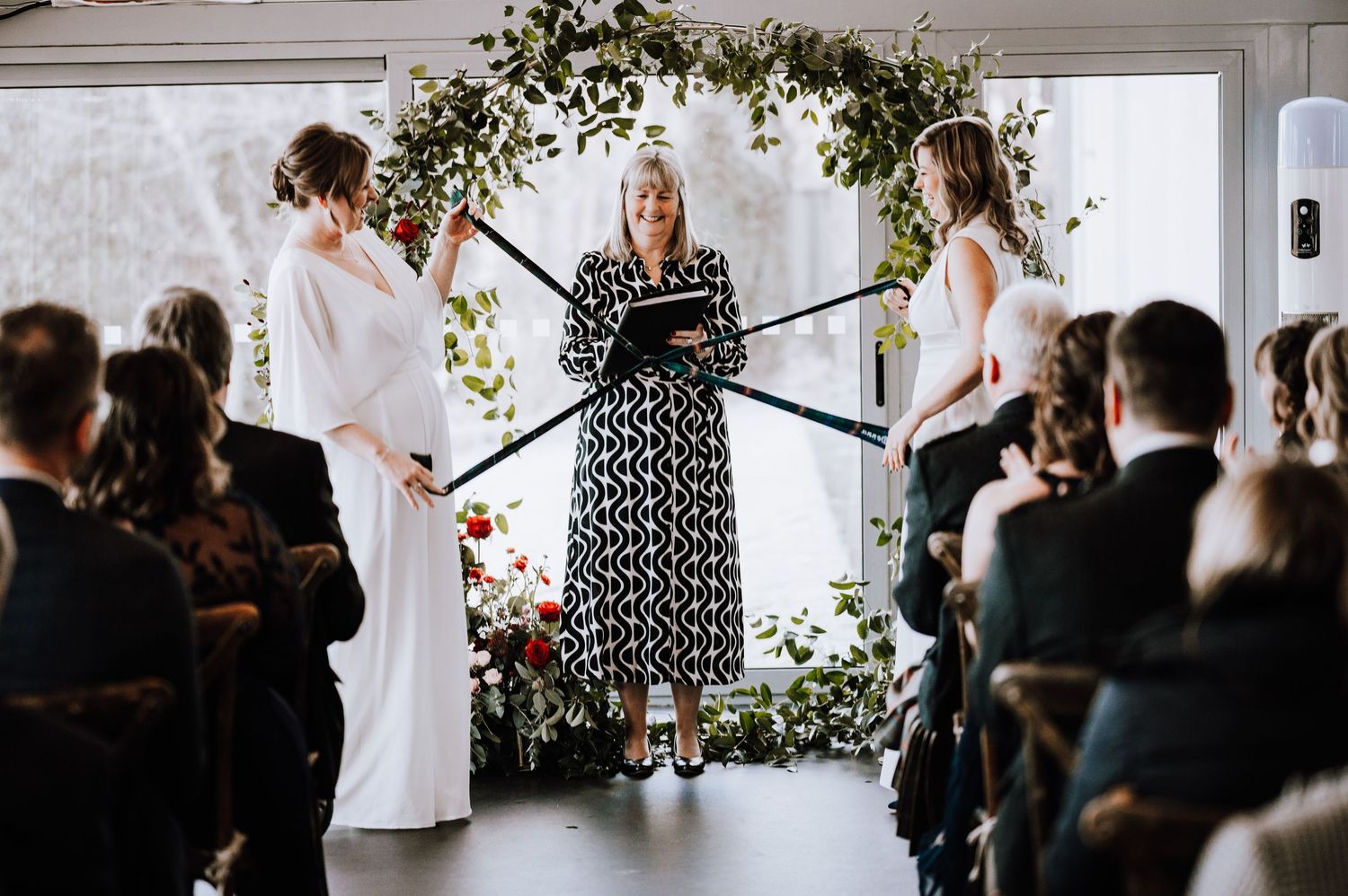 Same Sex Wedding Photography. Indoor wedding ceremony setup with greenery arch and guests seated in rows.