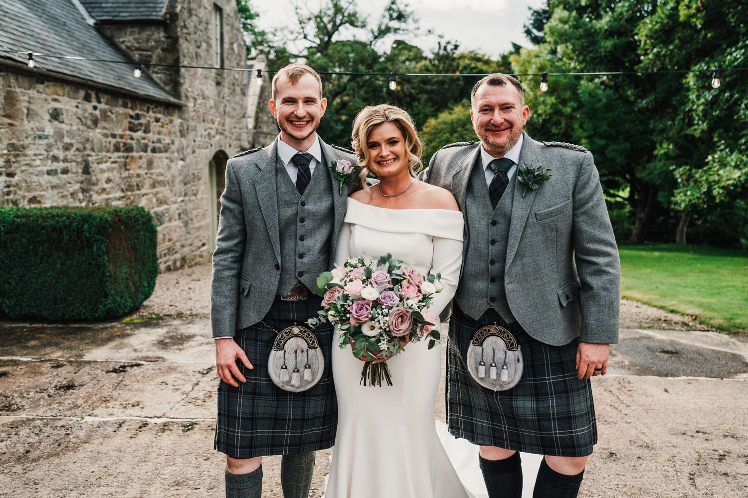 Wedding party in traditional Scottish kilts poses together outside a stone building.