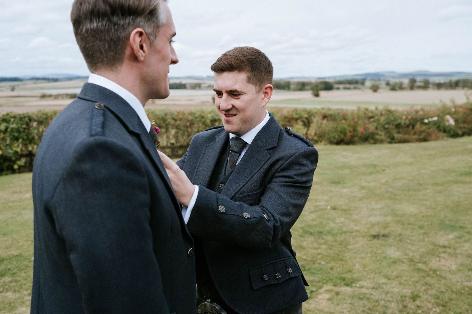 Wedding party members in formal attire chatting on a grassy hillside.