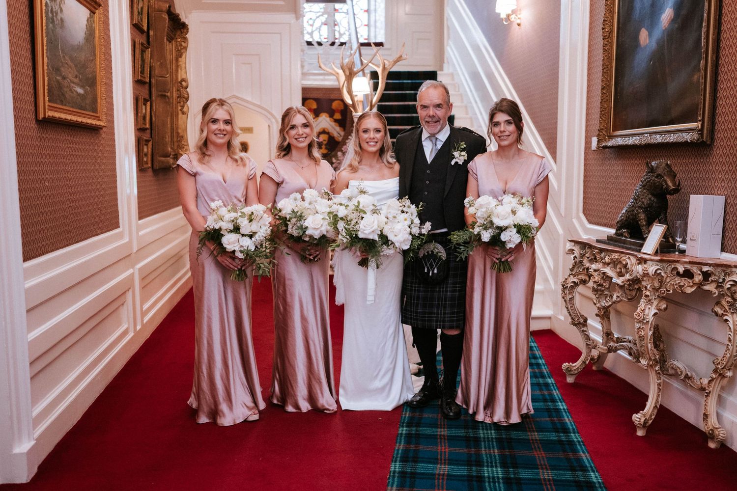 Wedding party lined up in an elegant pink-walled room with ornate decor and chandeliers.