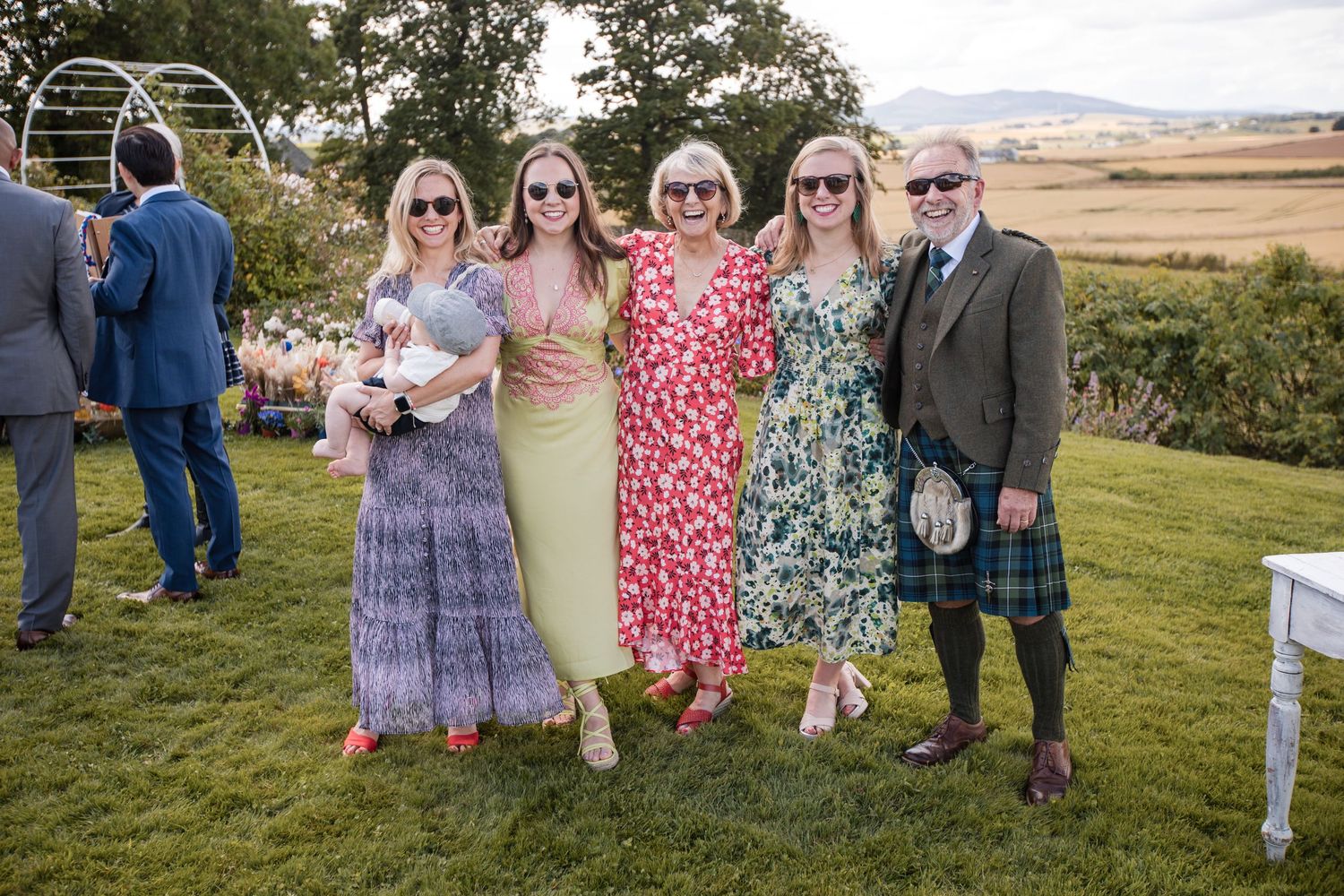 Wedding guests in colorful dresses gather on a grassy lawn for outdoor photos.