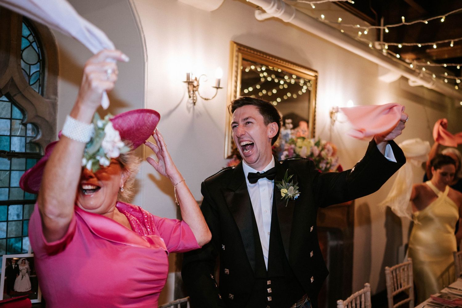 Guests celebrating with raised arms under twinkling lights at an elegant indoor wedding reception.