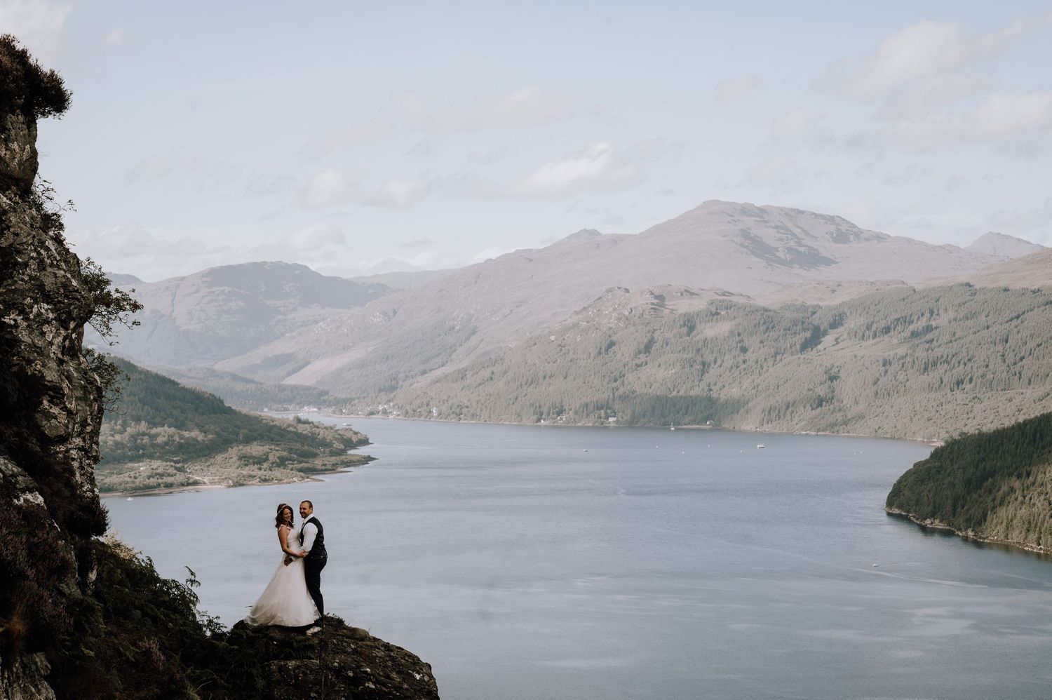 Distant silhouettes of newlyweds pose on a cliff overlooking a majestic mountain lake.