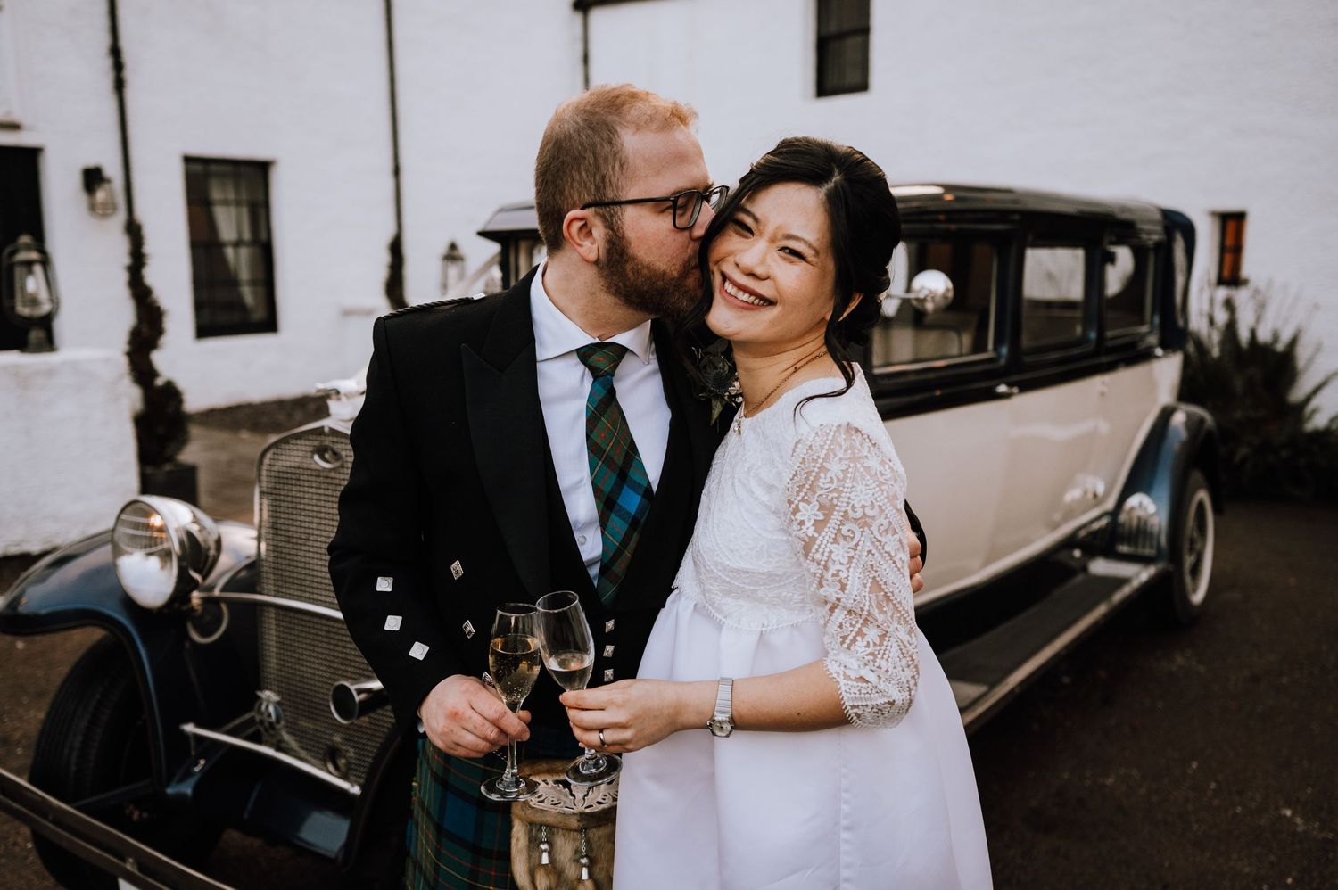 Candid moment of laughter shared between newlyweds next to a vintage automobile.