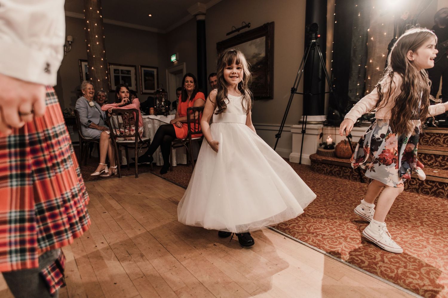 Children in formal wear twirling on a wooden dance floor at a Scottish wedding reception.
