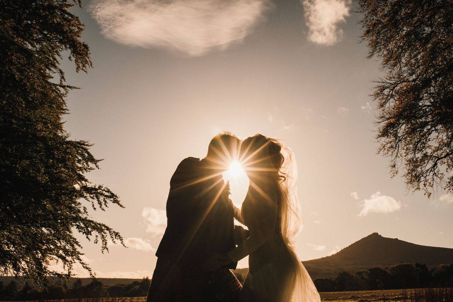 Silhouette of newlyweds kissing with sunburst through trees and mountain backdrop at sunset.