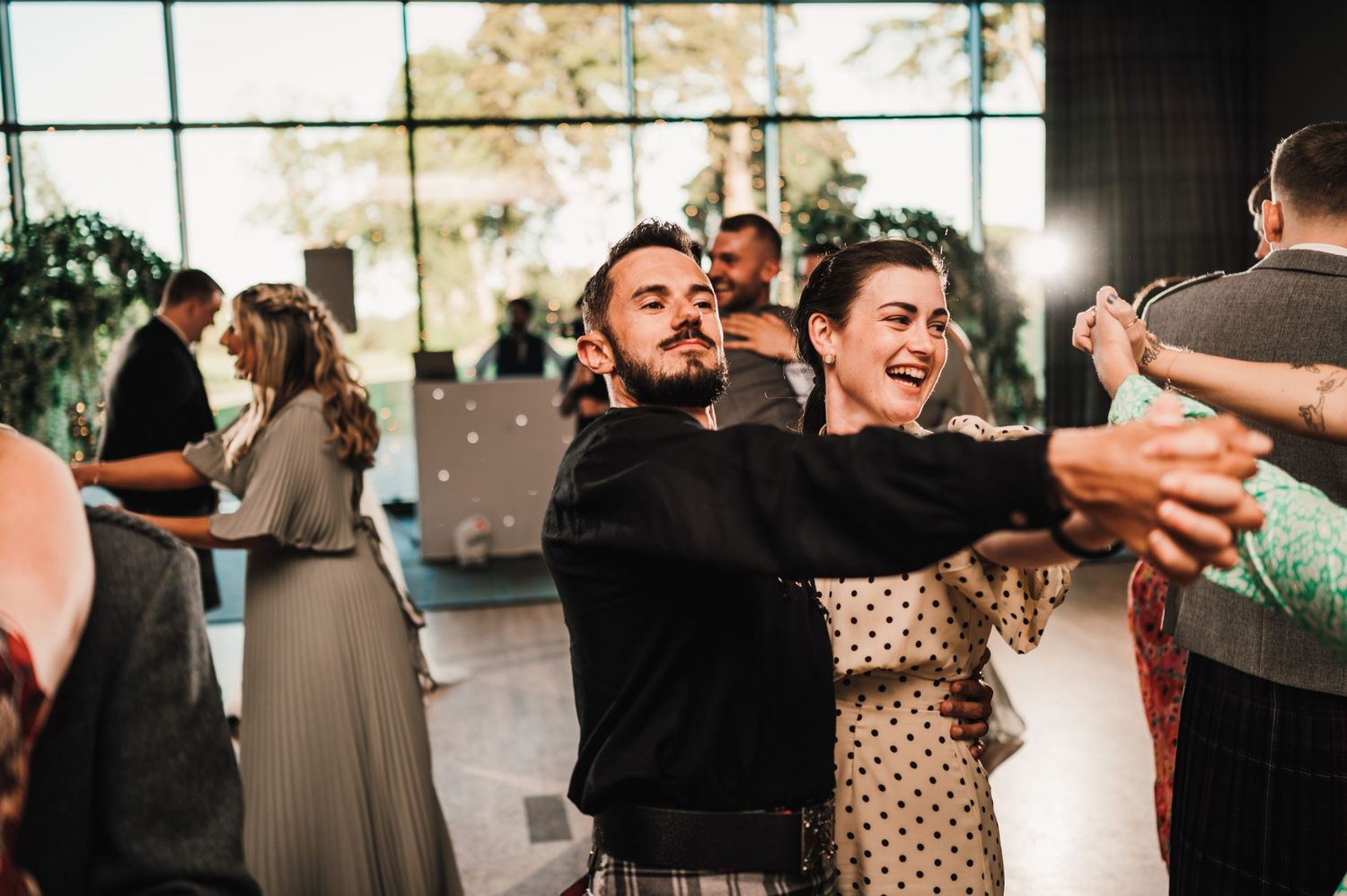 Wedding guests enjoying dancing and laughing at a reception in a venue with large windows.