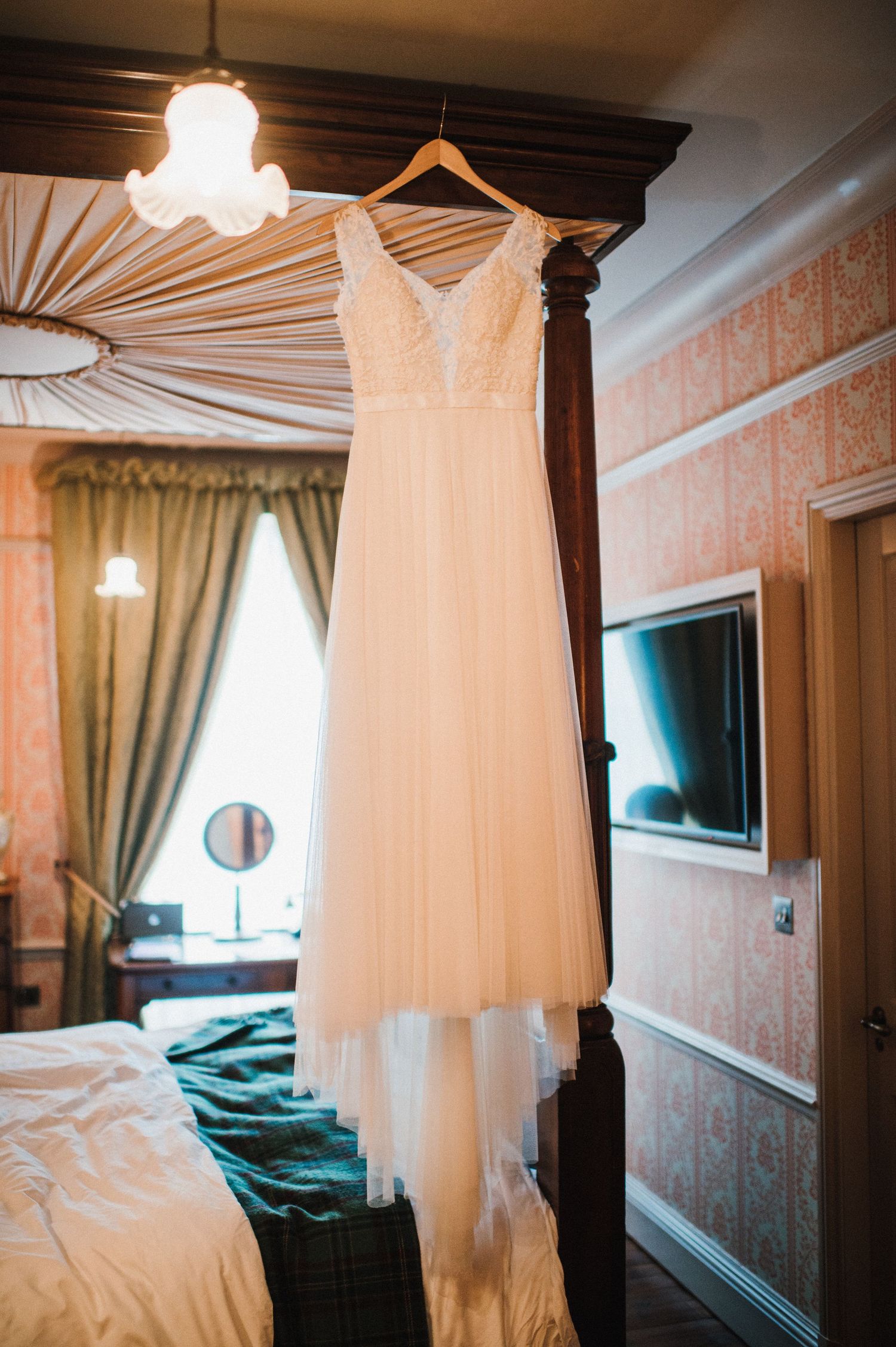 An elegant white wedding dress with lace details hanging from a four-poster bed frame.