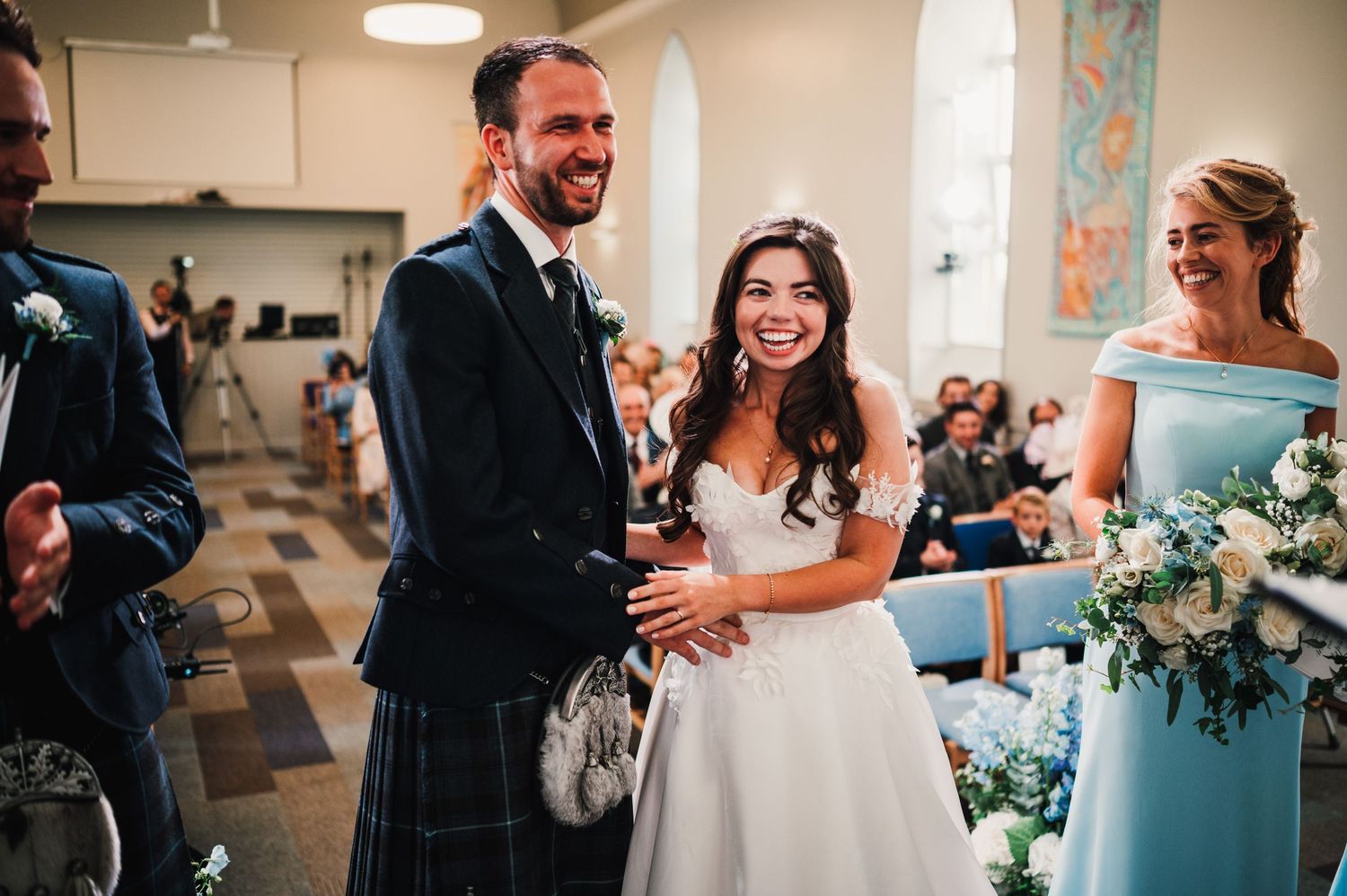 A wedding ceremony filled with joy and laughter as attendees celebrate in a church hall with blue floral decorations.