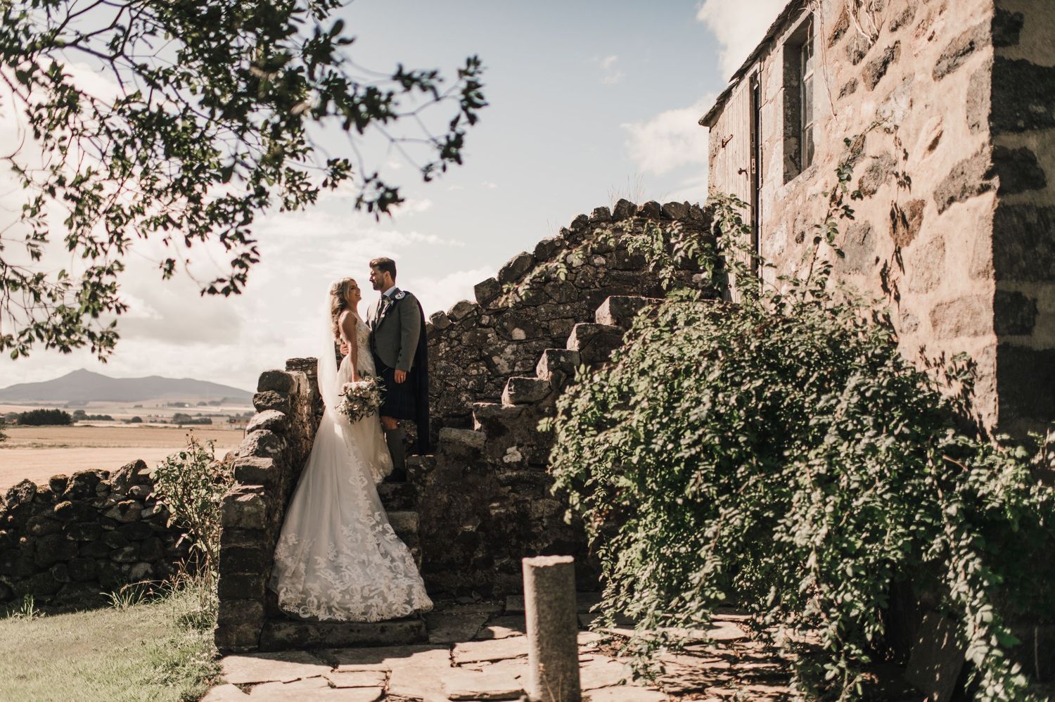 Wedding couple stands on stone steps with scenic countryside views.