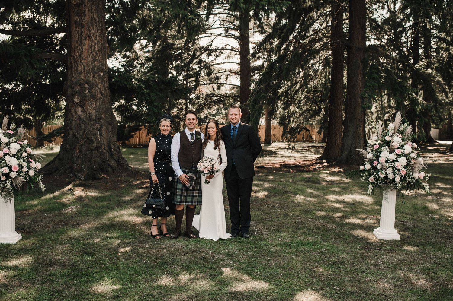 Wedding group portrait with Scottish kilts in front of towering evergreen trees.