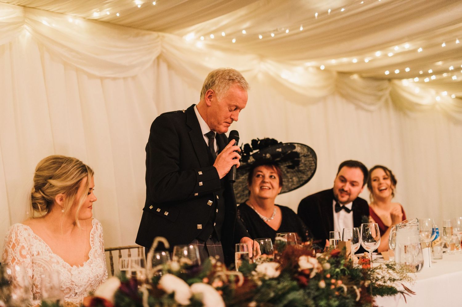 Wedding reception speech being delivered at an elegant head table decorated with floral arrangements.