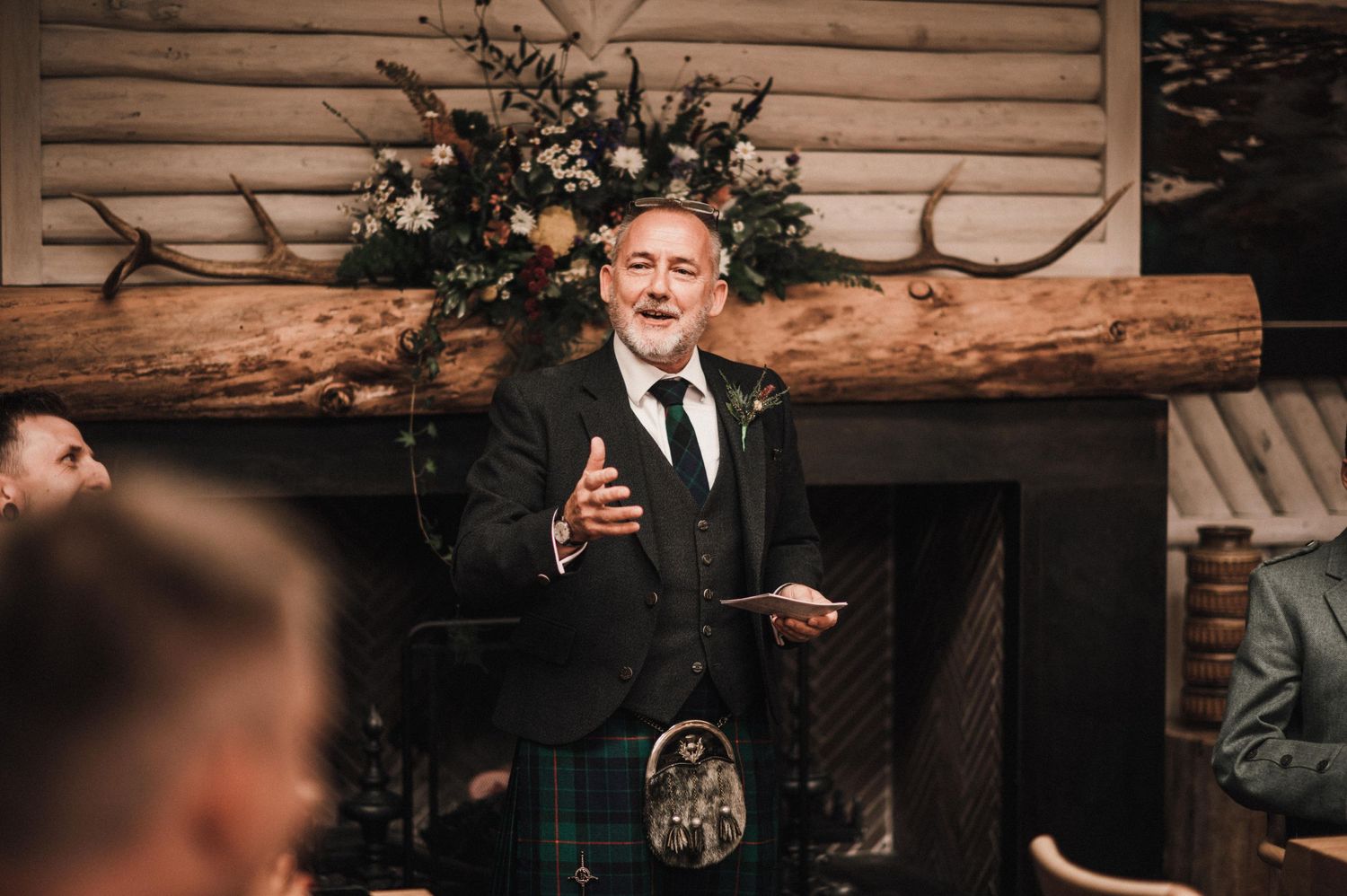 Speaker at podium during celebration event with rustic decor and floral arrangements.