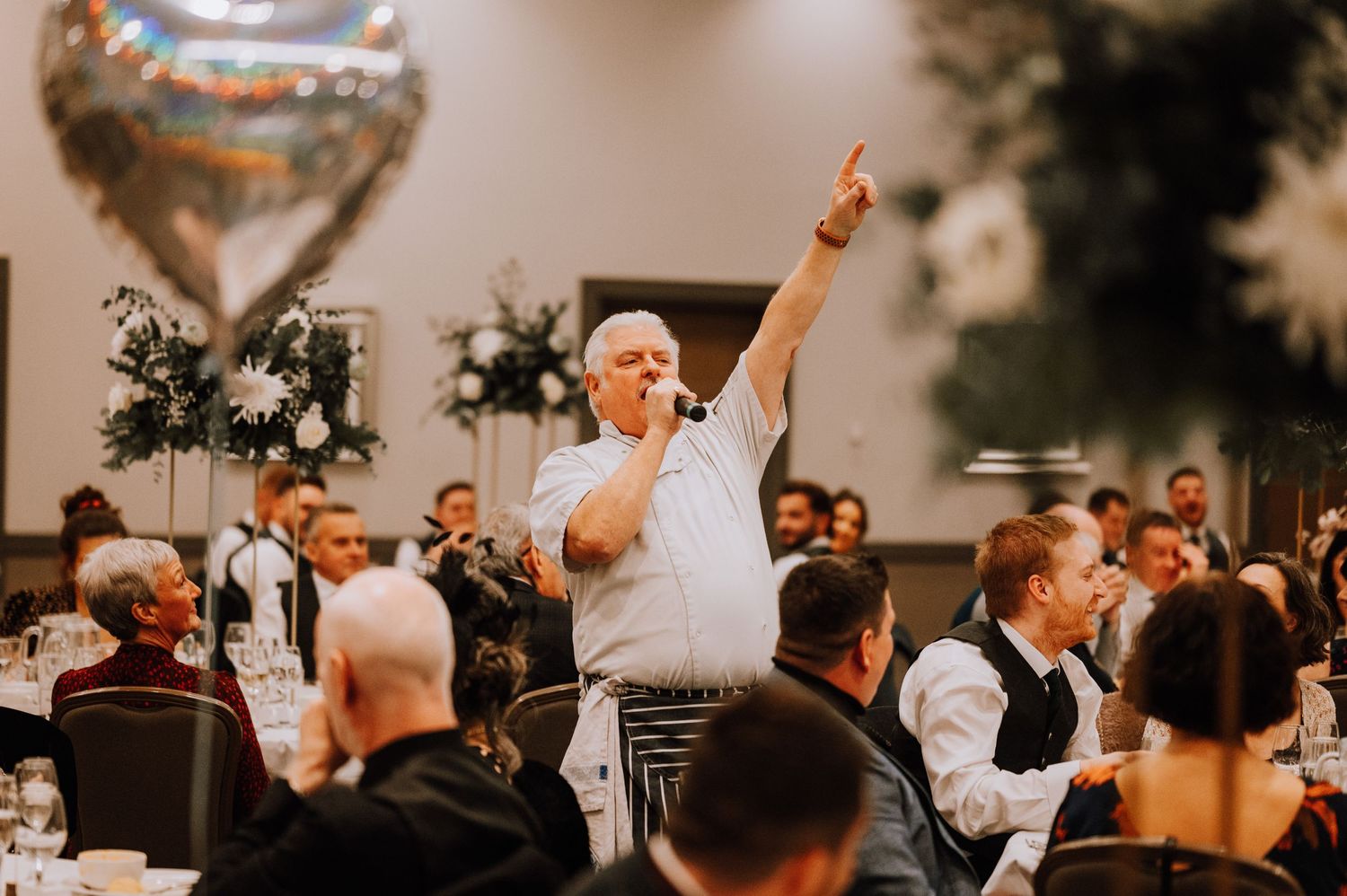 Guest stands enthusiastically during wedding reception celebration with decorative balloons and floral arrangements.