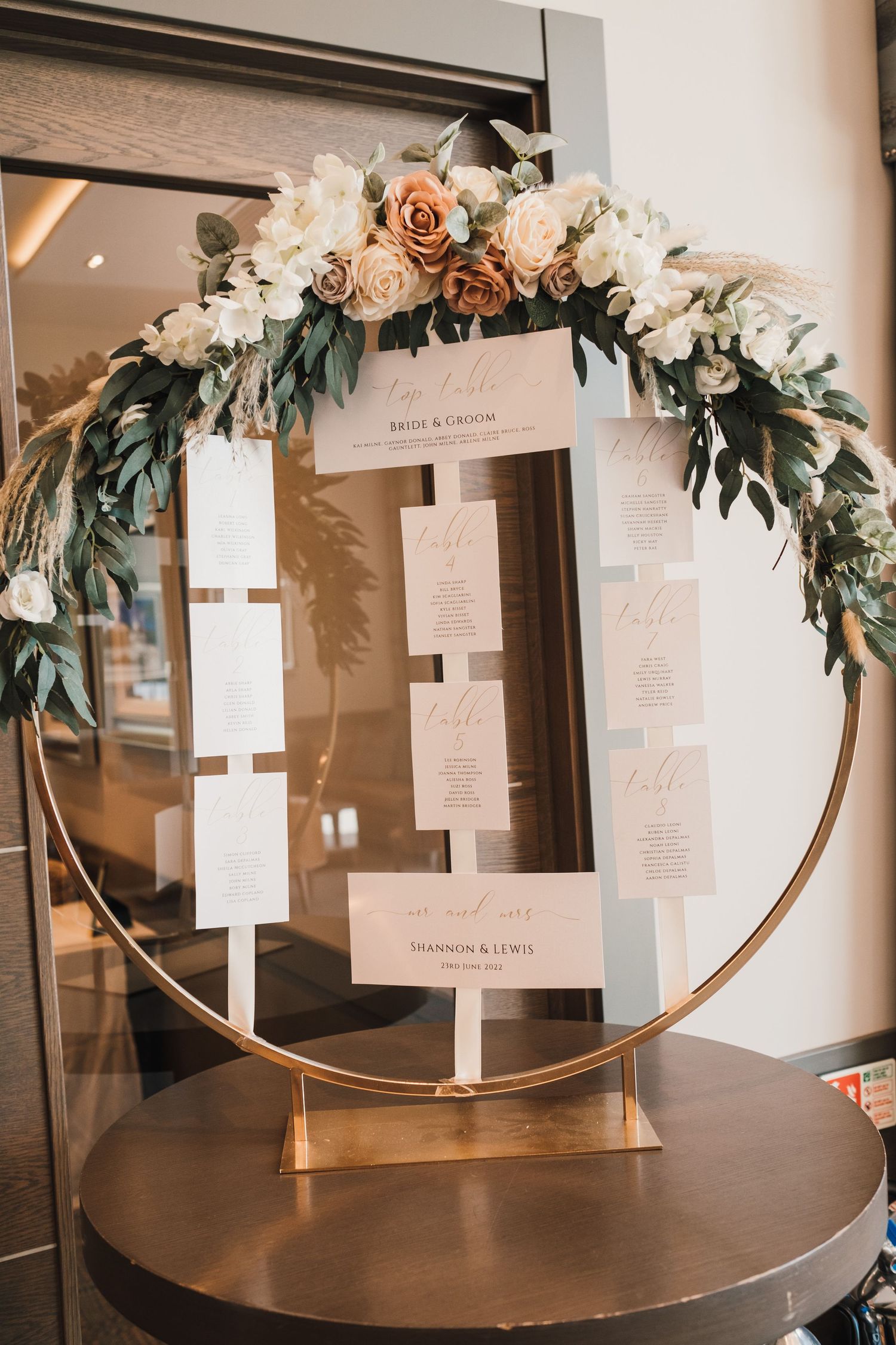 Circular gold hoop wedding seating chart decorated with blush roses and greenery.