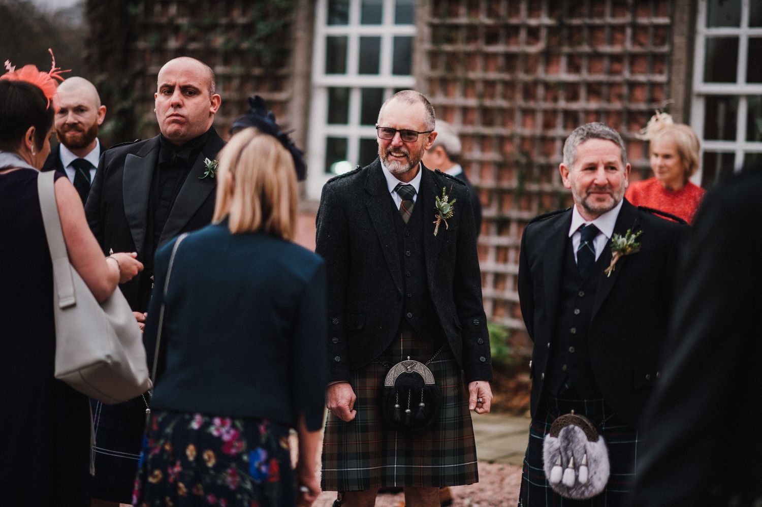 Wedding guests in kilts and formal attire mingle outside historic brick building.