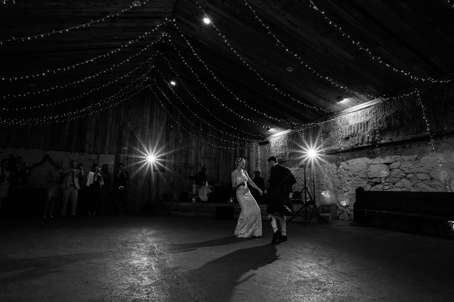 A black and white photo captures a wedding dance beneath twinkling fairy lights in a rustic venue.