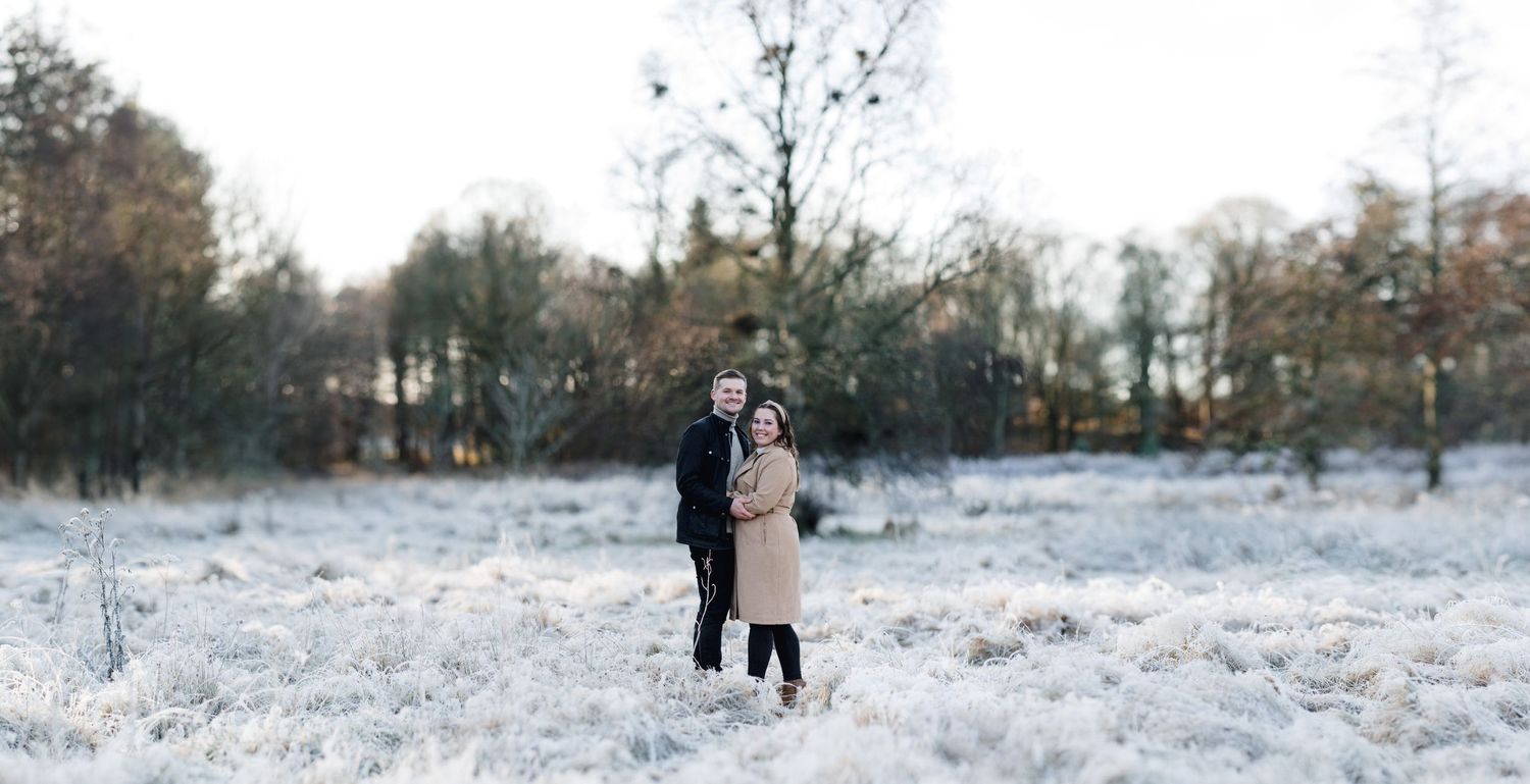A couple embraces in a snowy field surrounded by winter trees at sunset.