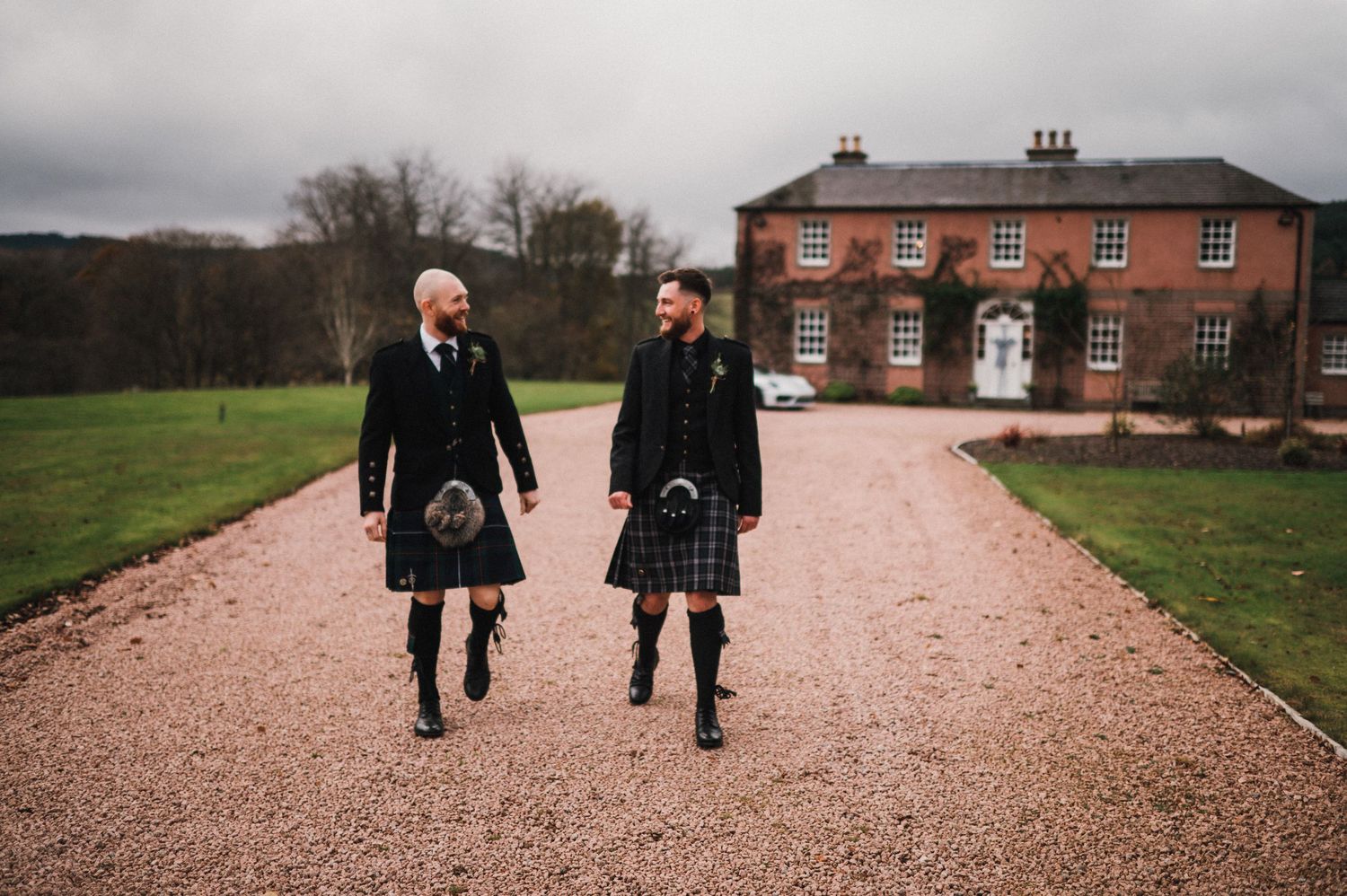 Two people in traditional Scottish kilts walk together on a gravel path leading to a stately brick manor house.